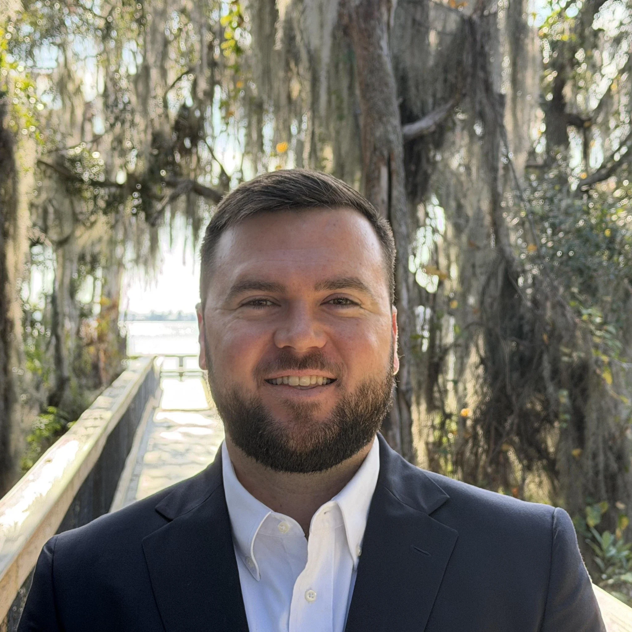 A man with short brown hair and a beard, dressed in a black suit and white shirt, smiling in front of a wooden bridge and trees.
