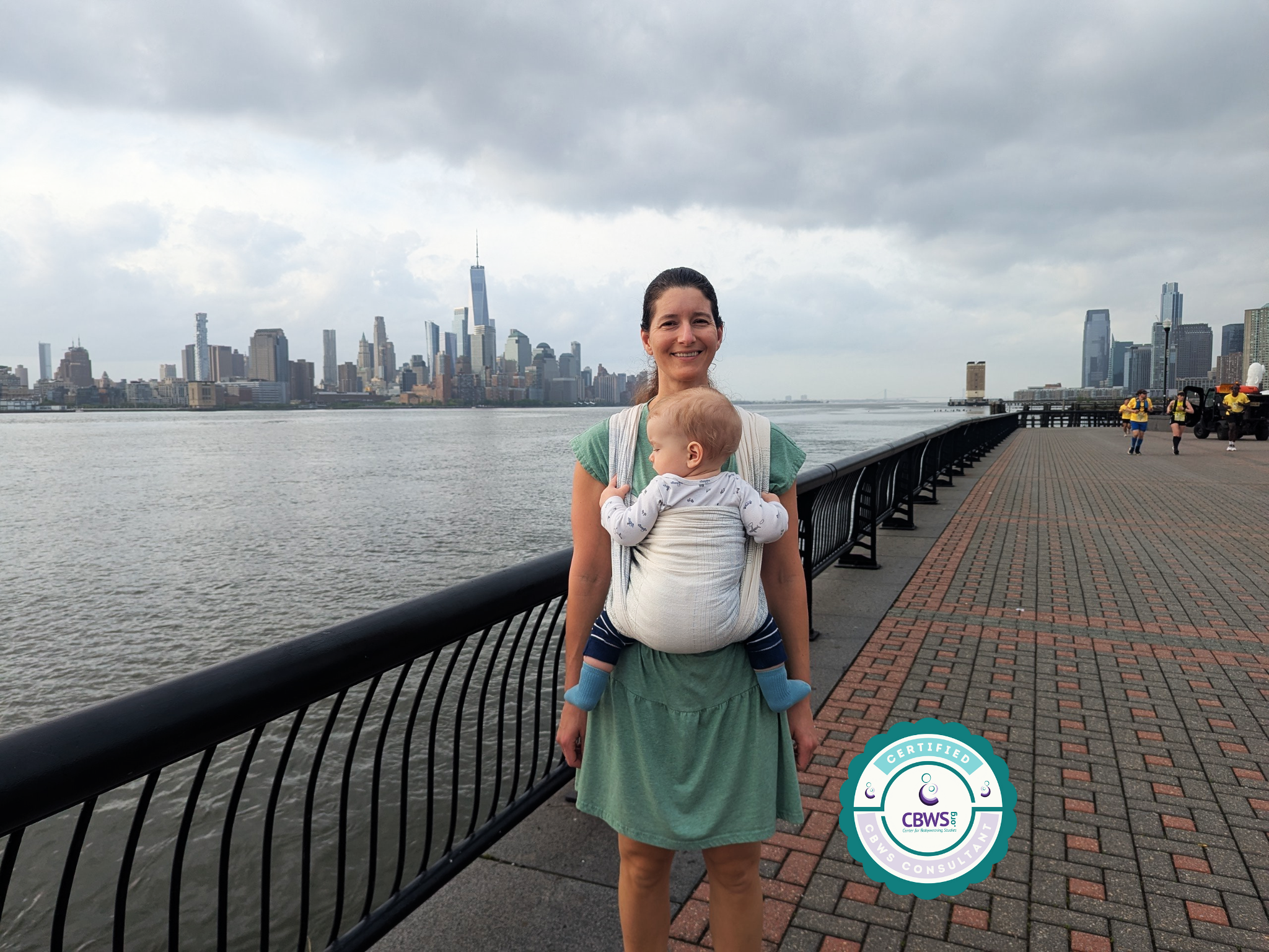 A smiling woman stands on a waterfront promenade with a baby in a woven wrap on her chest, overlooking a city skyline with tall skyscrapers under a cloudy sky.