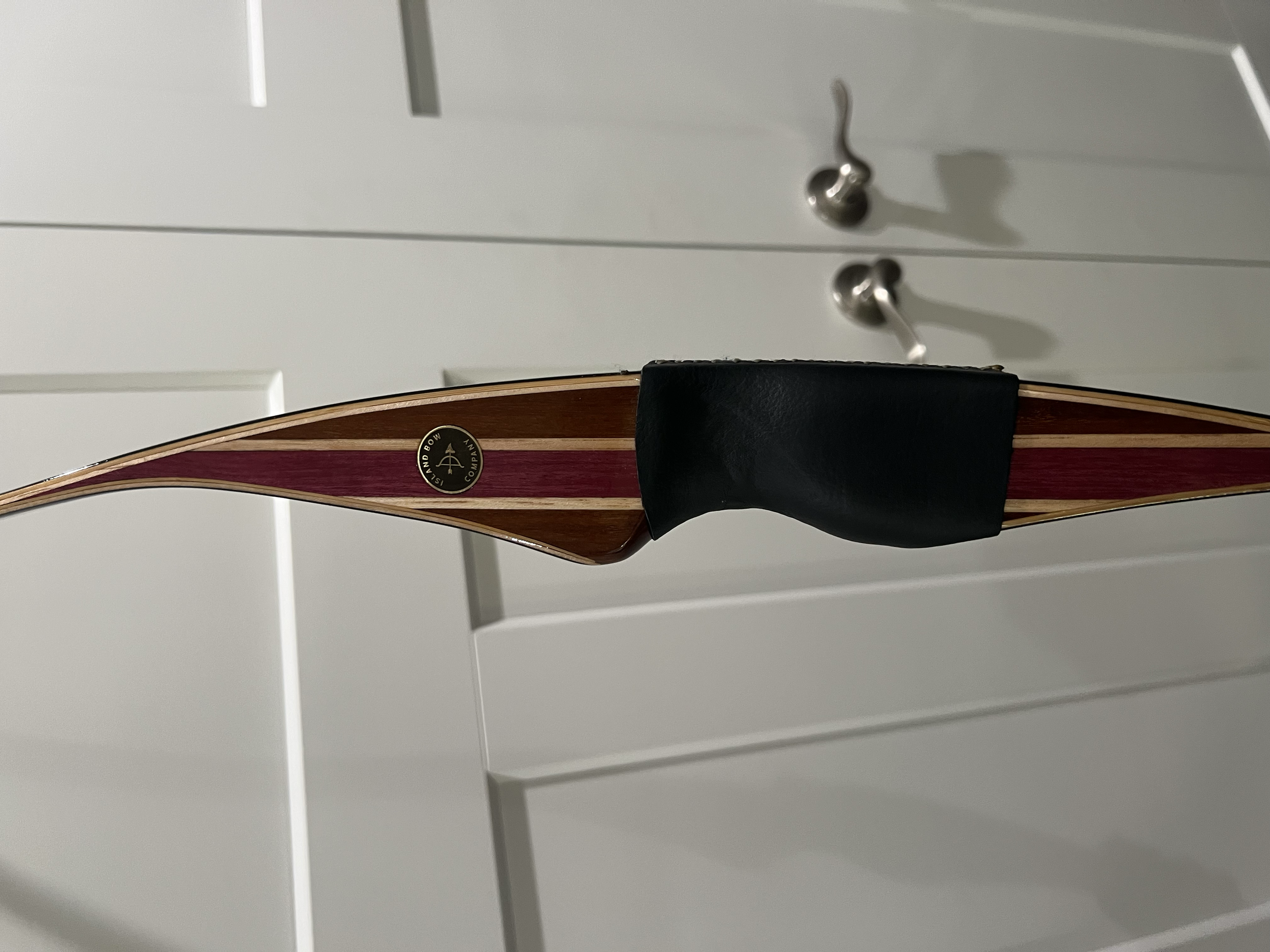 A close-up of a wooden bow with red and brown stripes, fitted with a black grip, against a white cabinet background.