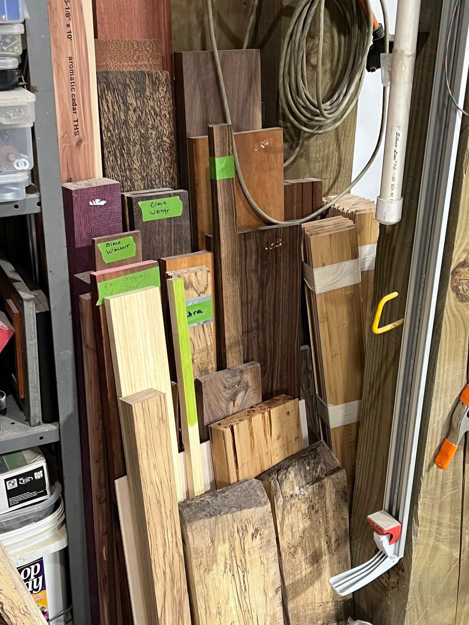 Various types of wood planks and boards stacked vertically in a workshop, with green labels indicating different wood types like black walnut, cherry, and oak.