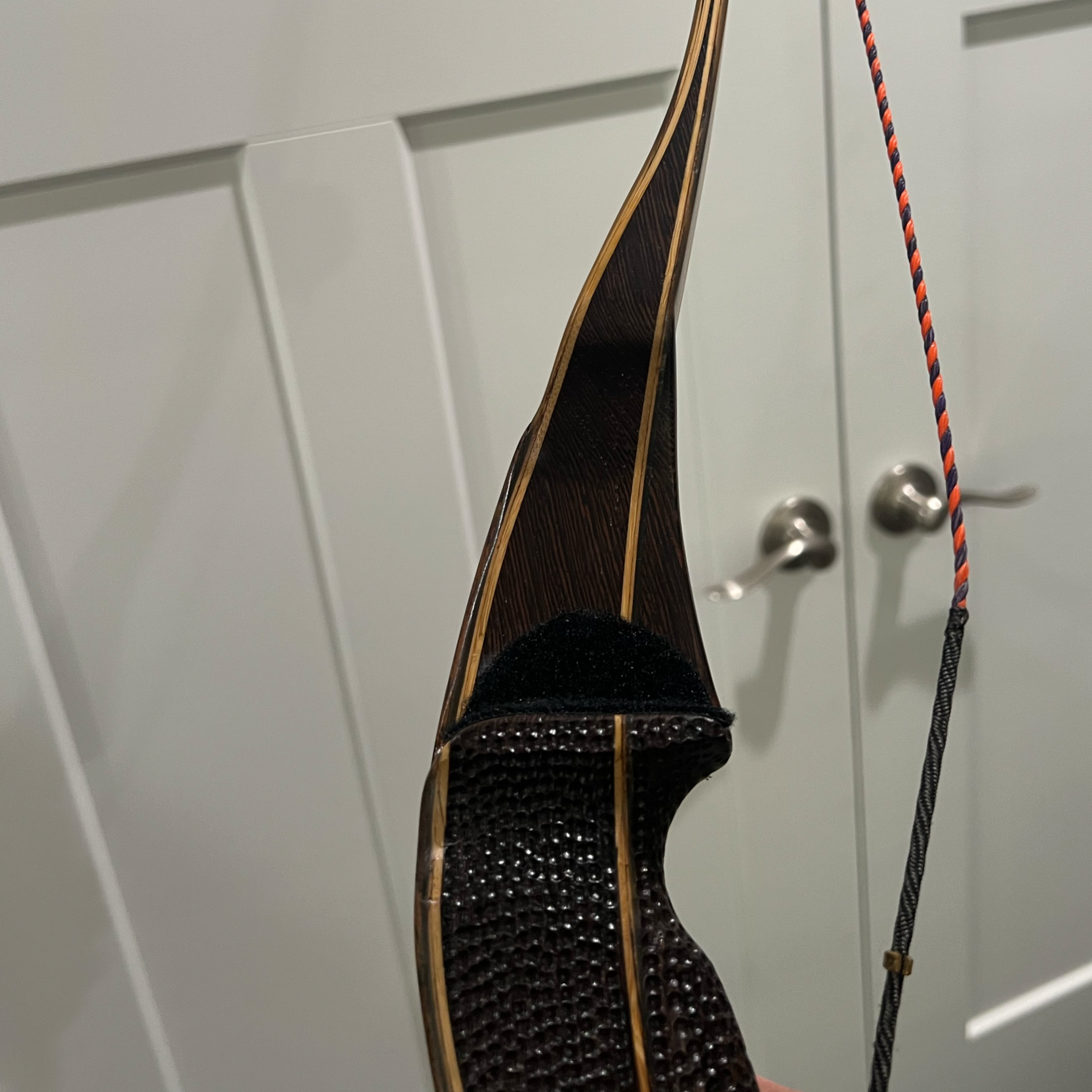 Close-up of a modern, curved wooden recurve bow with a textured black grip, positioned vertically against a white closet door with silver handles.