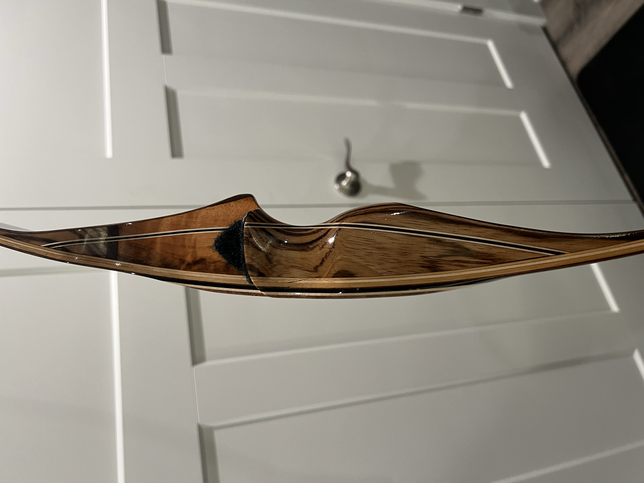 Close-up of a wooden recurve bow with a black arrow rest, facing a white cabinet with a metallic door handle.