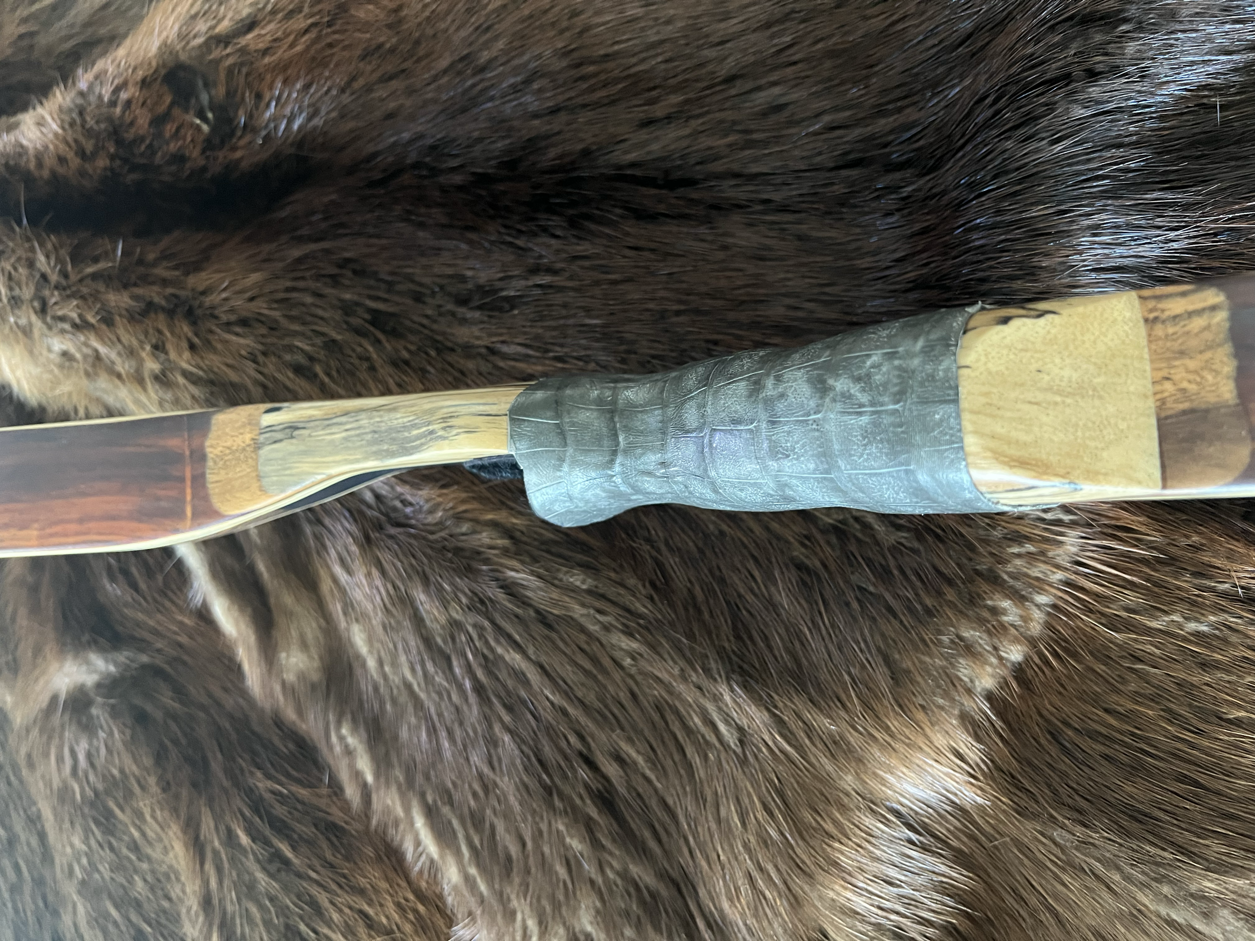Close-up of a hunting knife with a black leather-wrapped handle, a shiny stainless steel blade, and a leather sheath, resting on a brown furry animal hide.