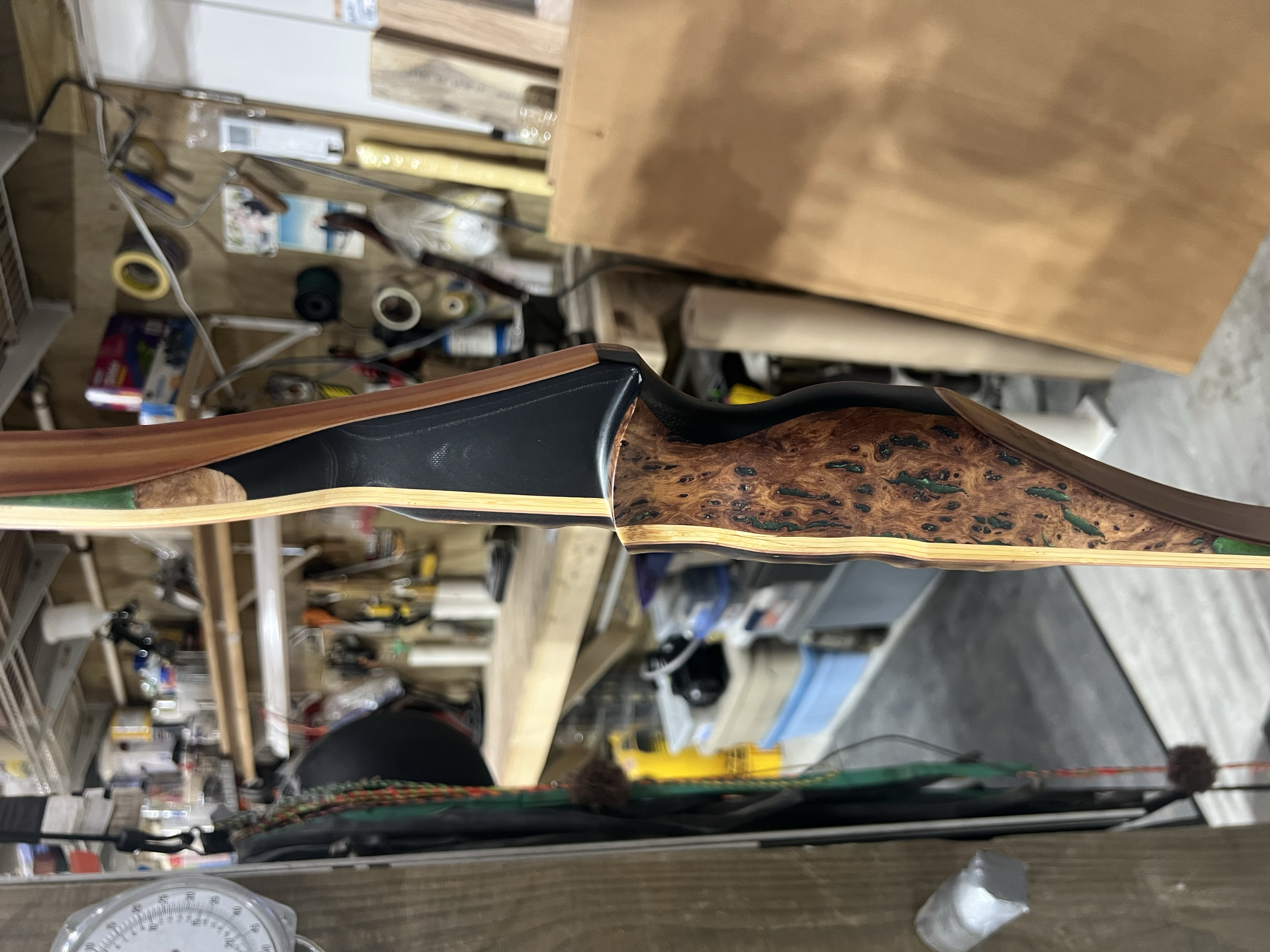 Close-up side view of a decorative bow with a glossy black finish and a wood grain pattern, resting on a workbench in a workshop.