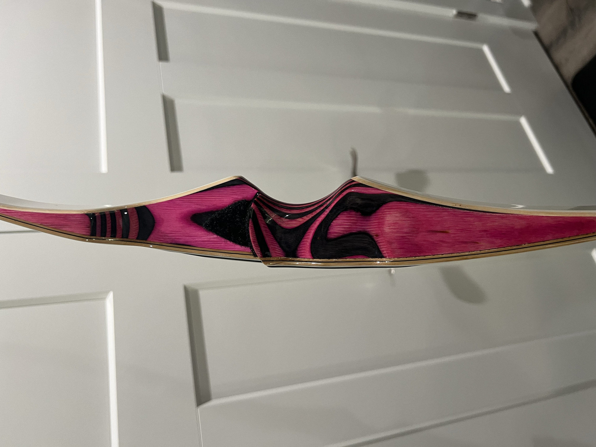A close-up of a bow for archery with pink and black swirling patterns, hanging in front of a white cabinet.