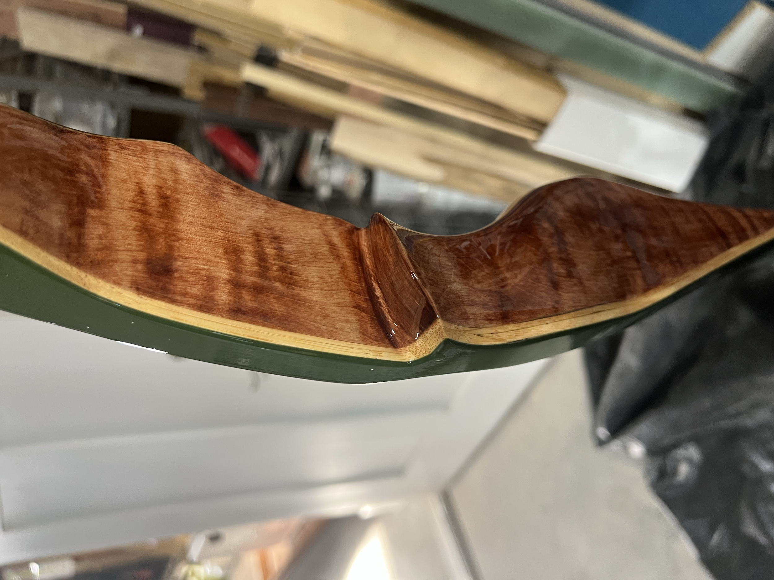 Close-up of the edge of a wooden guitar body showing its glossy finish and smooth curves, with a background of various wood planks and workshop items.