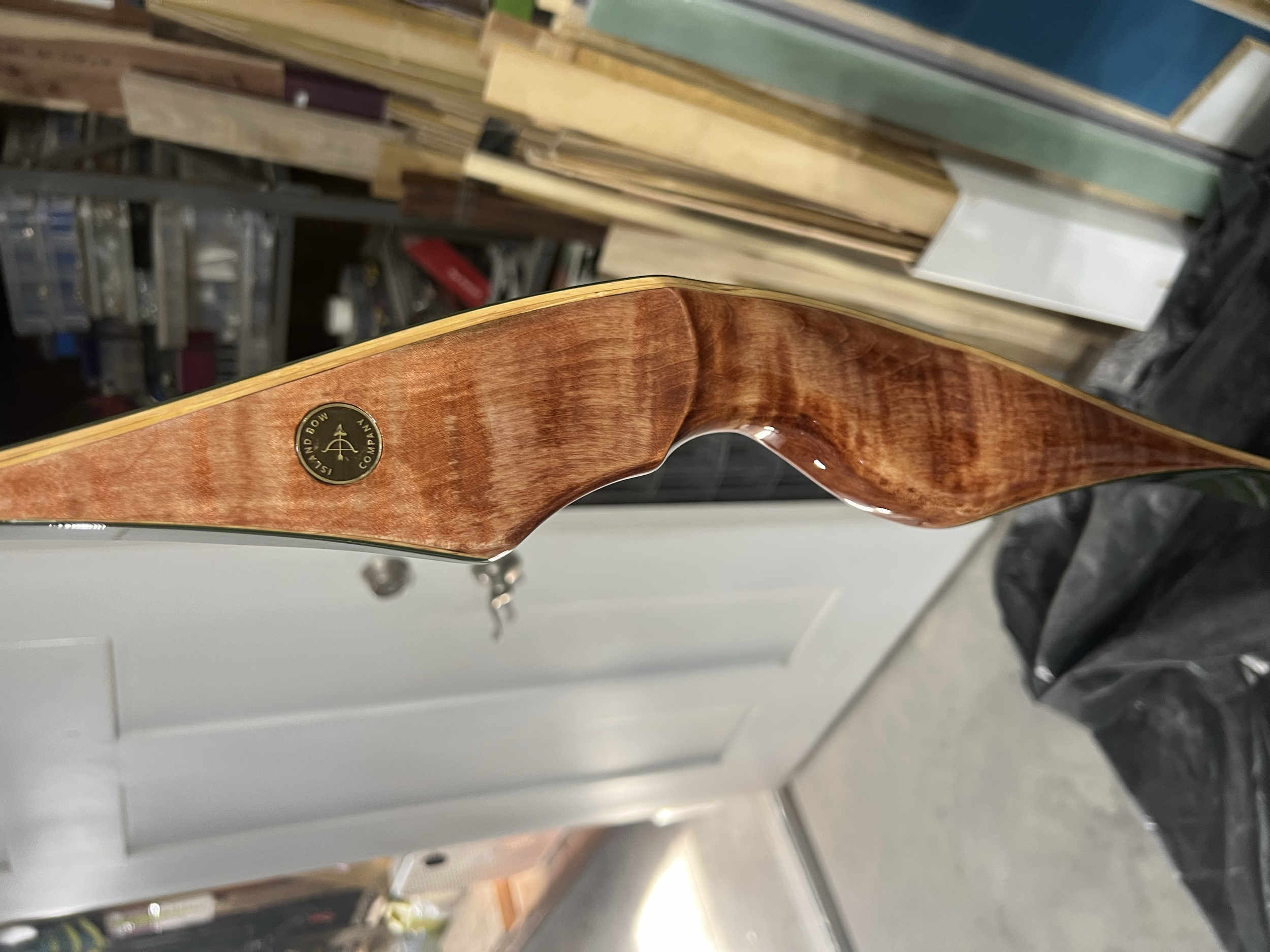 Close-up of the back of a wooden archery bow with a round black and gold sticker, in a workshop with wood and tools in the background.