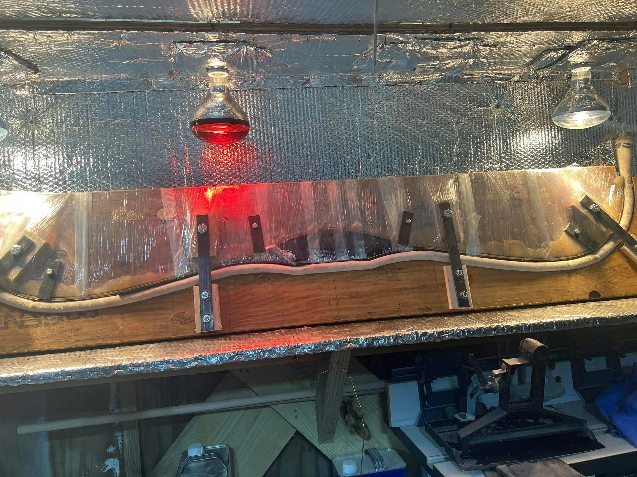 A science experiment setup with a curved transparent plastic shield, metal brackets, wooden base, fluid-filled glass containers, and heating elements, all on a metallic surface with a reflective insulation material in the background.
