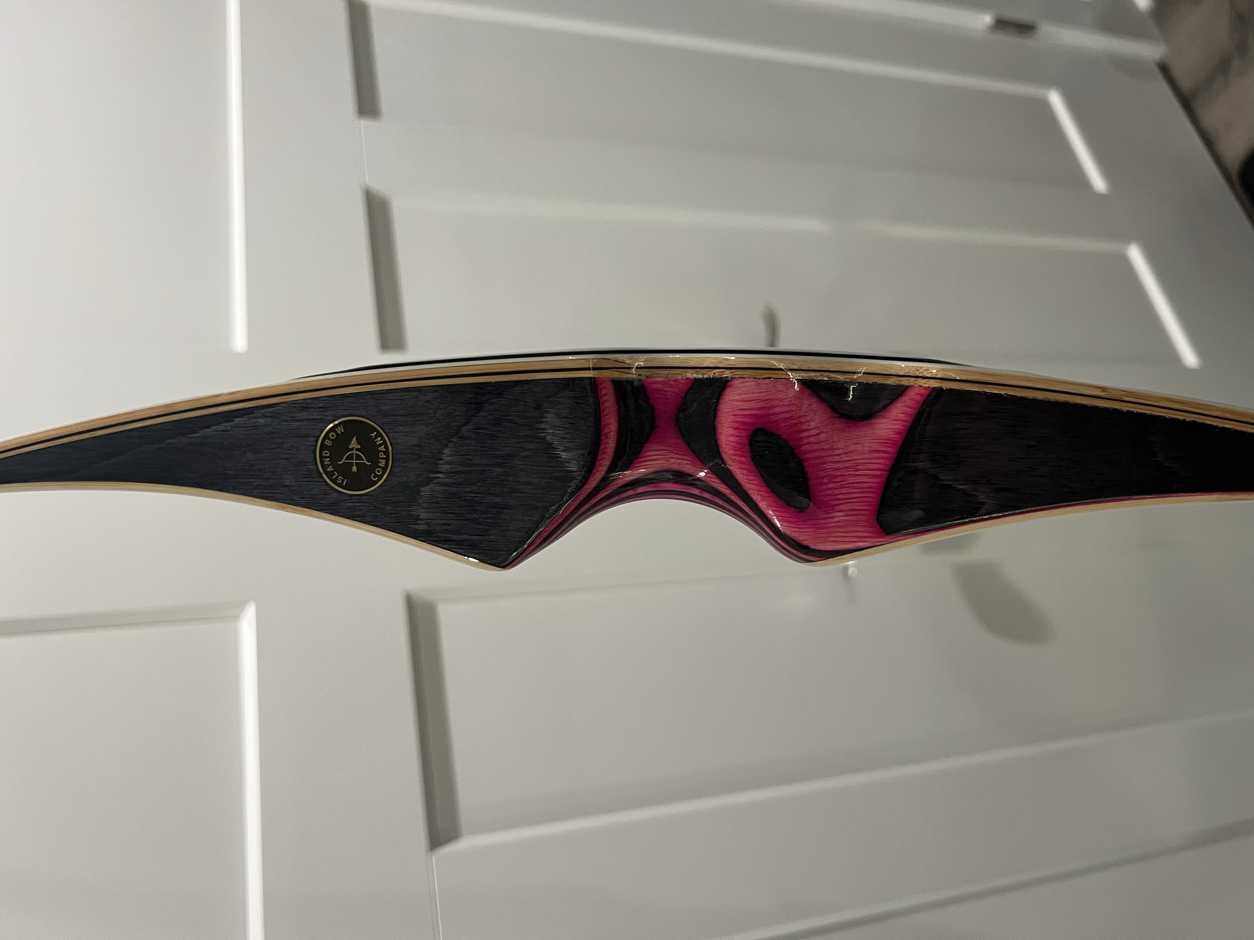 A close-up of a wooden recurve bow with black and pink accents, showing the grip and limb. The background features white cabinet doors.