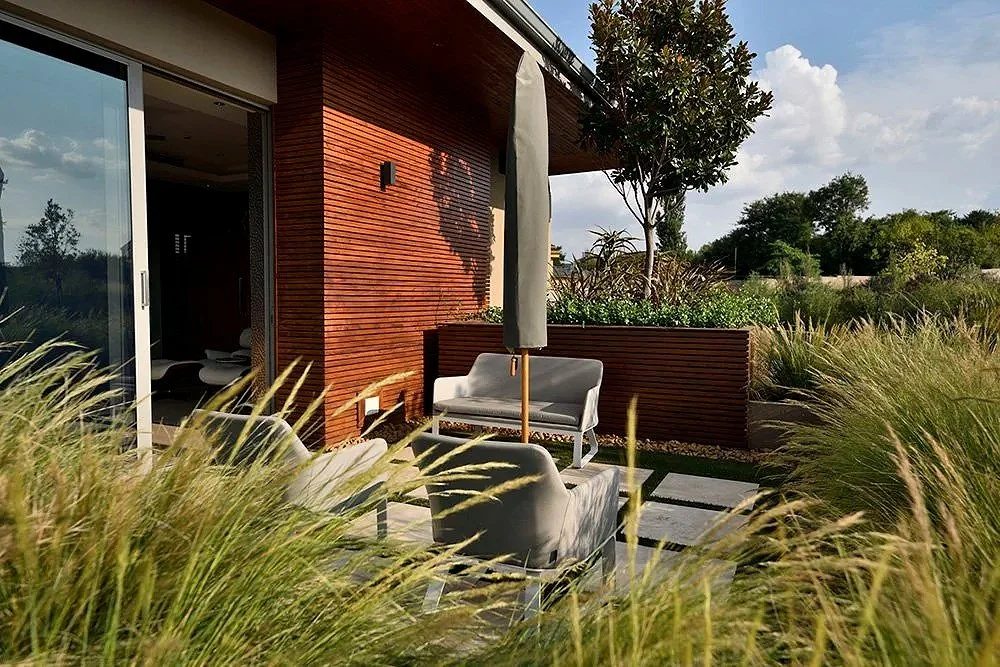 Outdoor patio with modern furniture, umbrella, and lush greenery next to a house with a wood exterior.