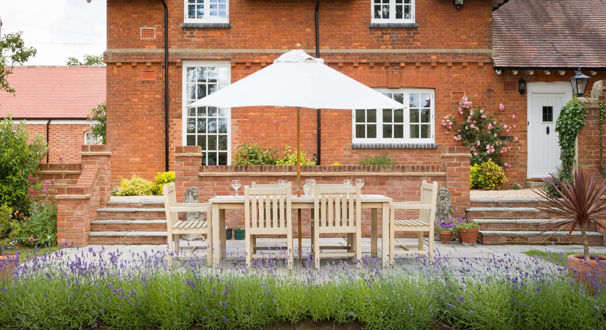 Brick house with garden patio, white outdoor dining table, several matching chairs, a large white umbrella, purple and pink flowering plants, potted plants, and brick steps leading to the house door.