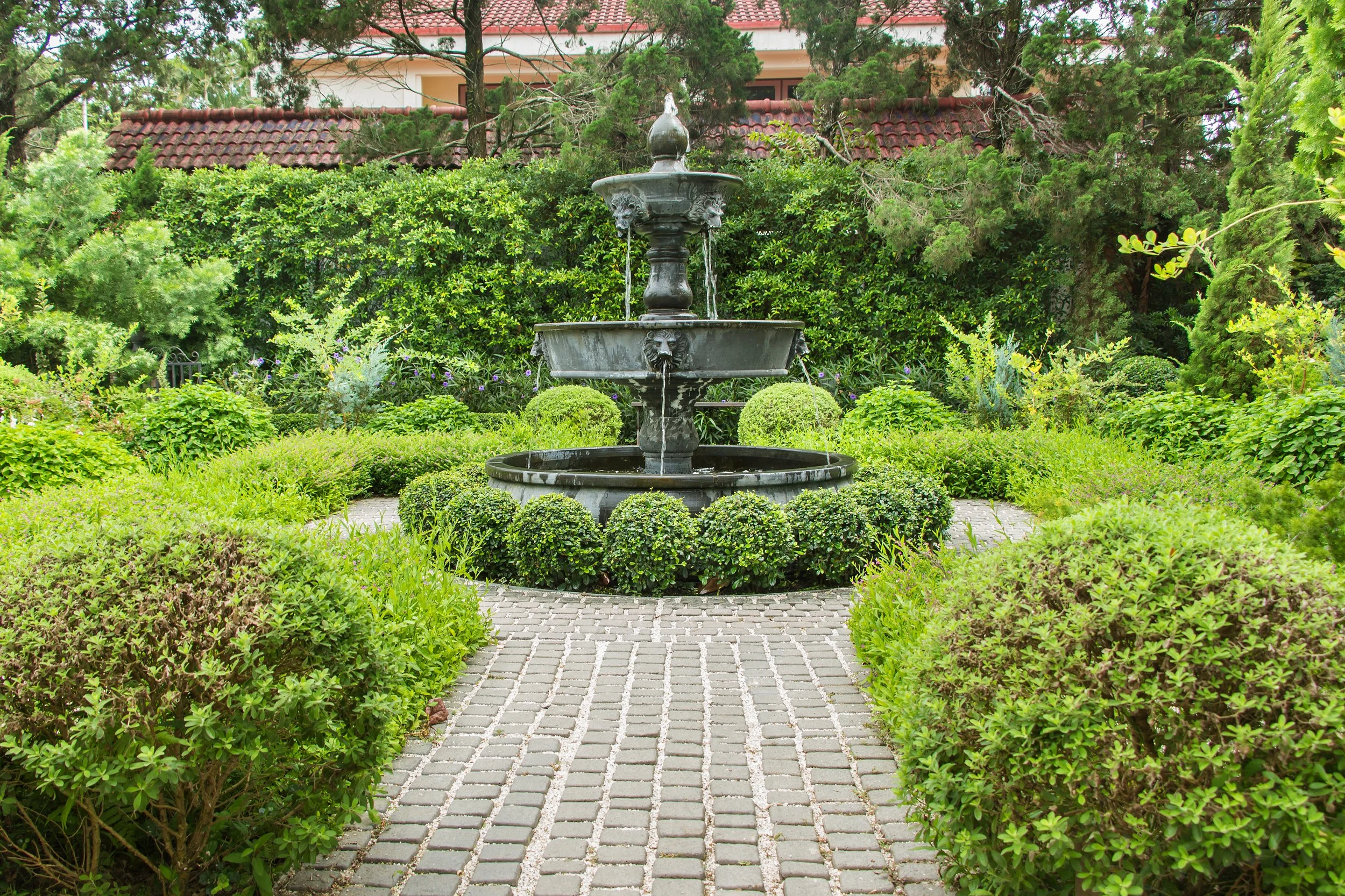 A black ornate fountain in the center of a lush green garden with various bushes and trees, paved pathway leading to it, and a house with a red-tiled roof in the background.