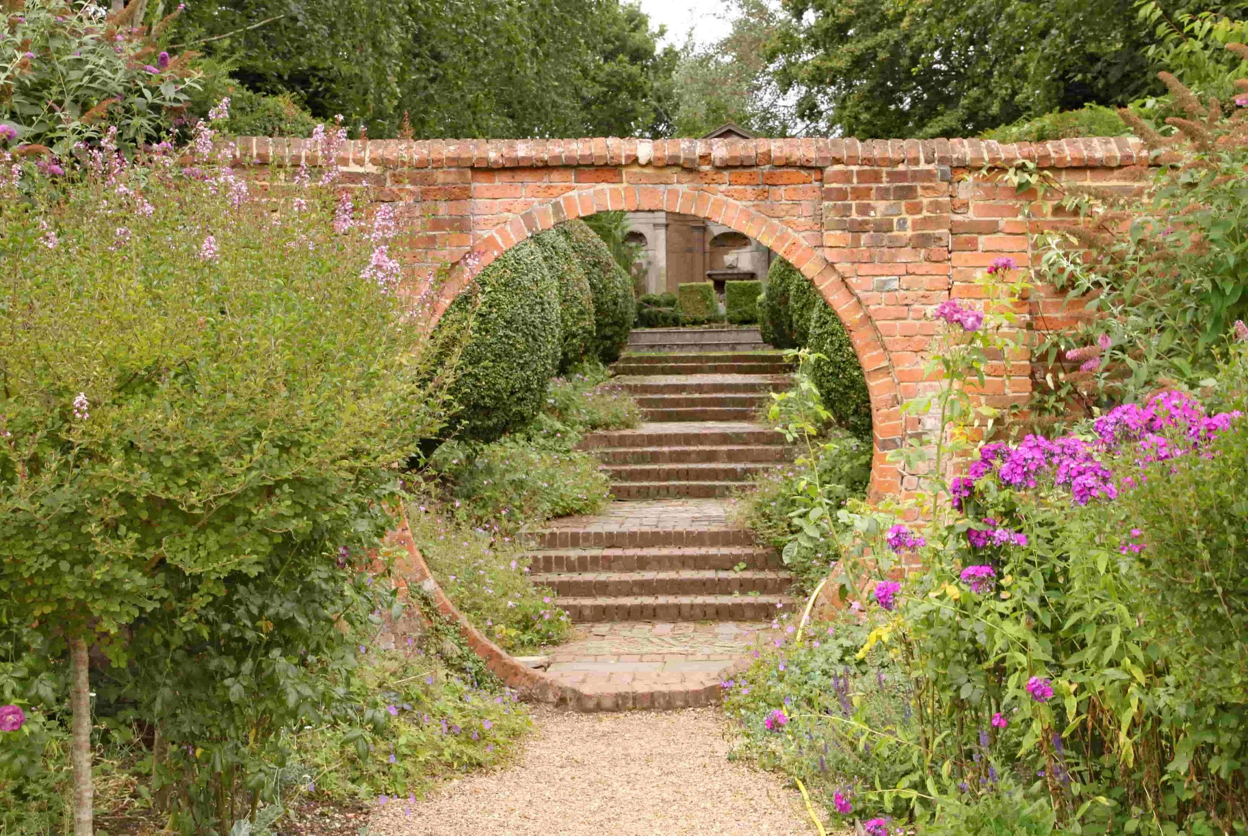 Clay brick wall with circular opening leading to stairs and topiary planting in a large Surrey garden