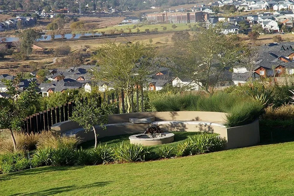 A landscaped backyard with a curved concrete seating area, a fire pit, green grass, trees, and other desert plants, overlooking a residential neighborhood with houses and a body of water in the background.