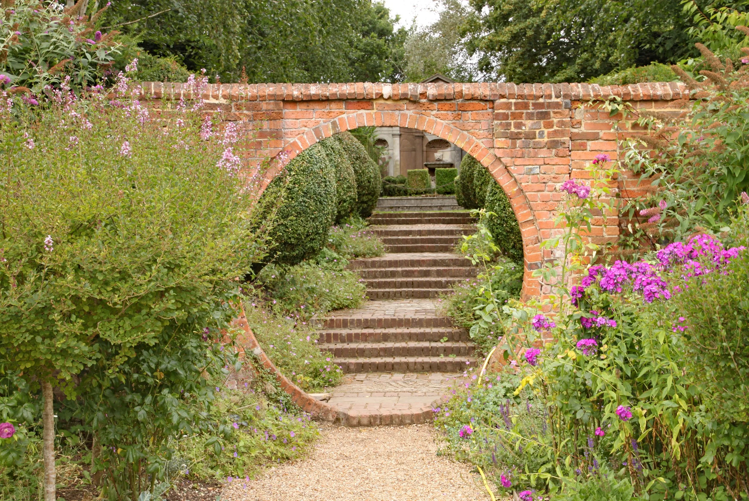 A garden with a brick archway opening to stairs leading to a fountain, surrounded by greenery and pink flowers.