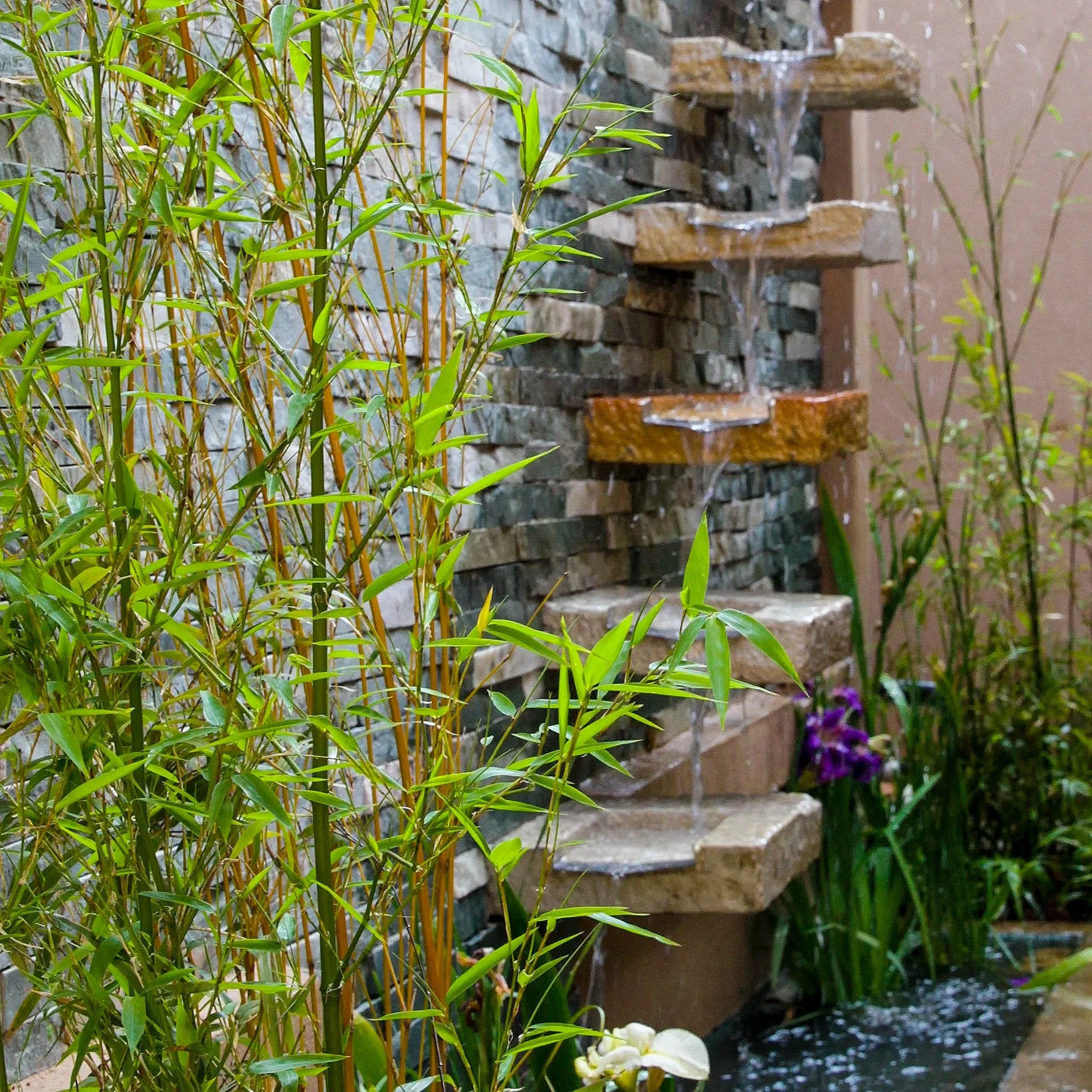 A cascading water fountain with water flowing over stone steps, surrounded by green plants and purple flowers.