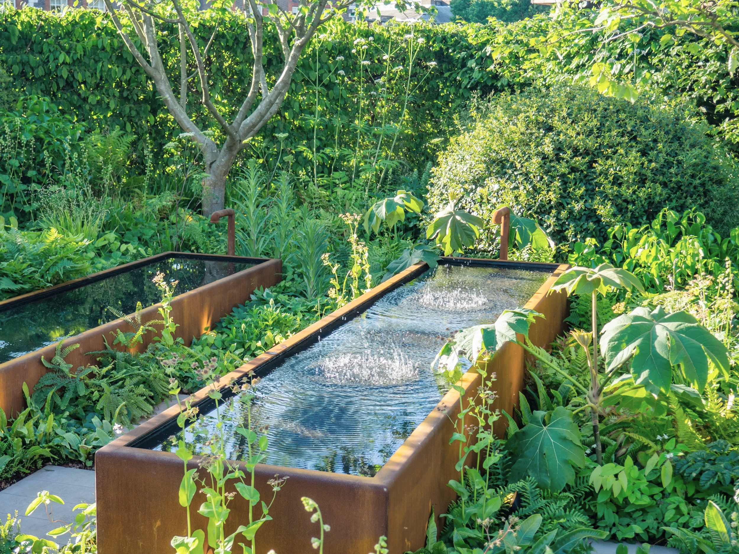 A lush, green garden with a variety of plants and trees surrounds two rectangular water features with fountains, made of dark metal, in the foreground.