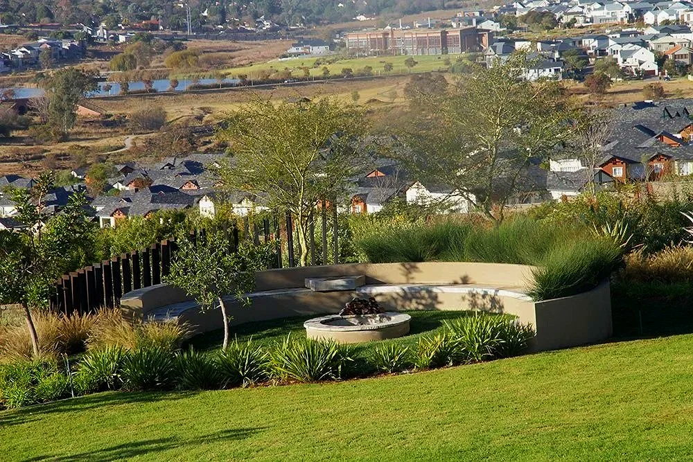A landscaped backyard with a curved seating area surrounding a fire pit, lush grass, and various plants, overlooking a neighborhood with houses and rolling hills in the distance.