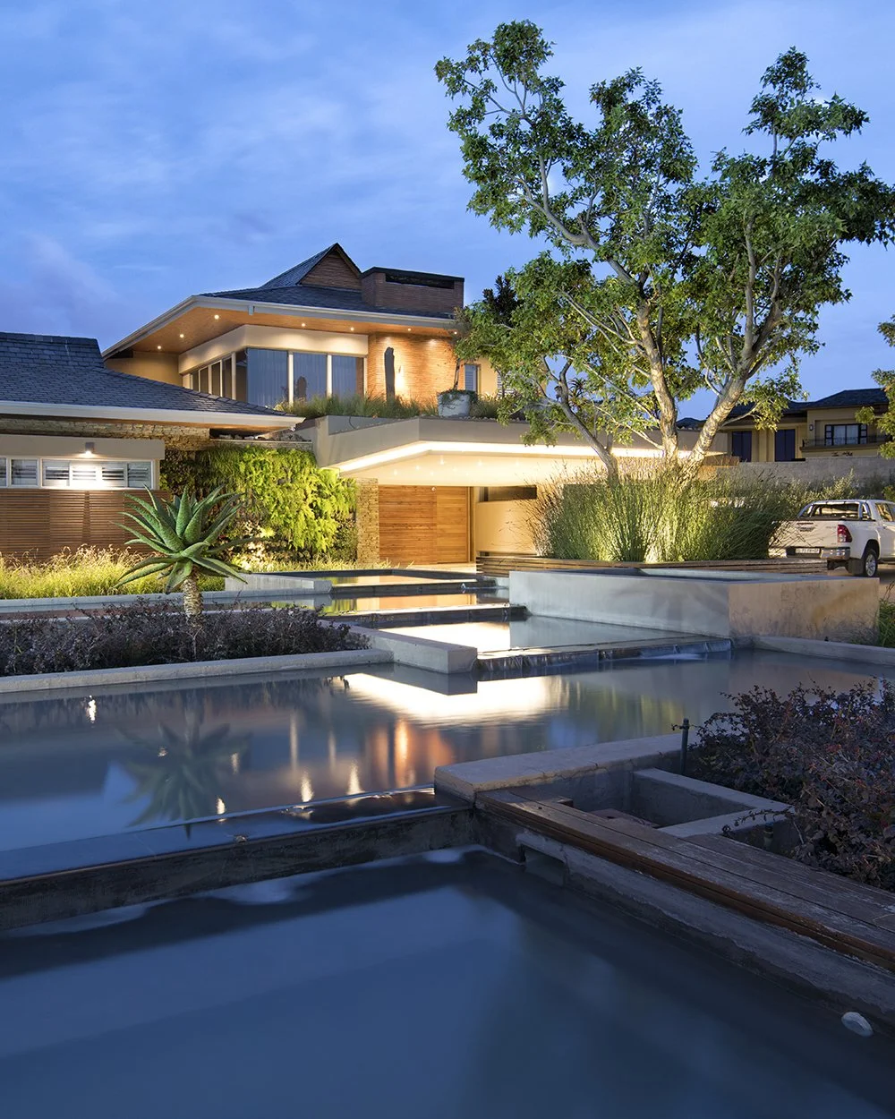 Modern house with outdoor water features, trees, plants, and a driveway with parked trucks during twilight evening.