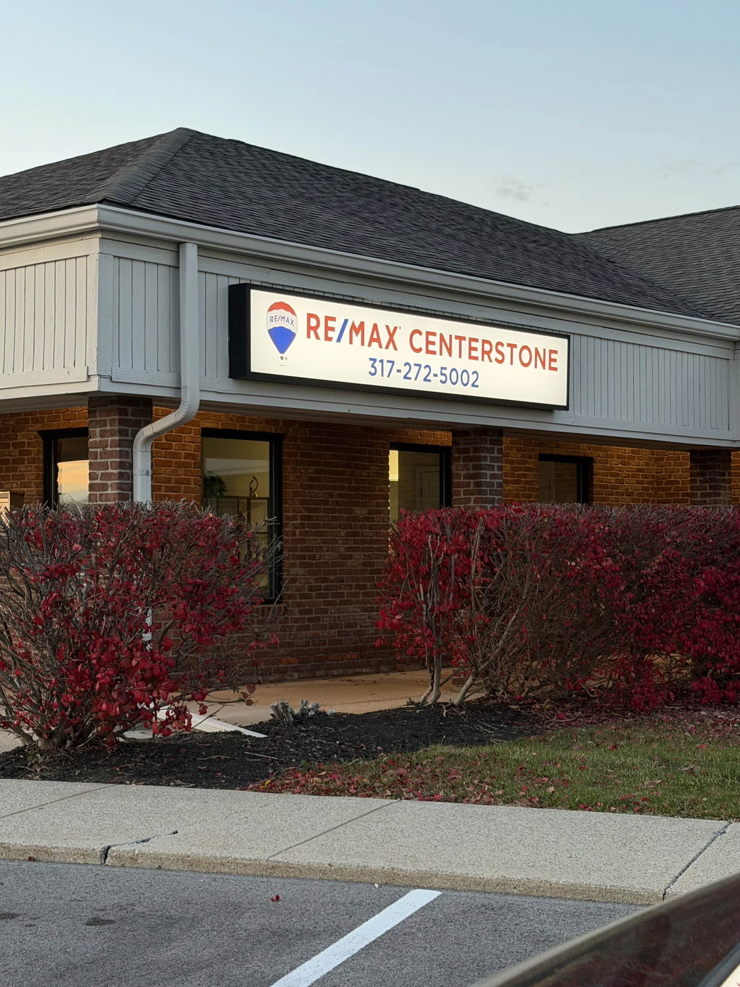 A brick building with a sign that reads RE/MAX Centerstone, a phone number, and the RE/MAX logo. There are red bushes and a sidewalk in front of the building.