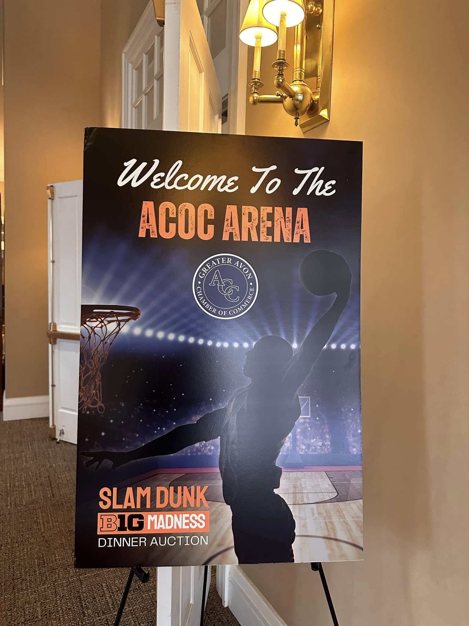 A basketball player silhouette dunking in a gym with a spotlight on their body and a basketball in their hand. The background shows a basketball court and appears to be an event poster for the ACoc Arena, featuring a dinner auction and a Slam Dunk event, with the Greater Avon Chamber of Commerce logo.
