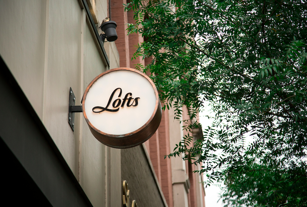 A round illuminated sign with the word "Lofts" in cursive font, hanging on the exterior of a building next to a tree with green leaves.