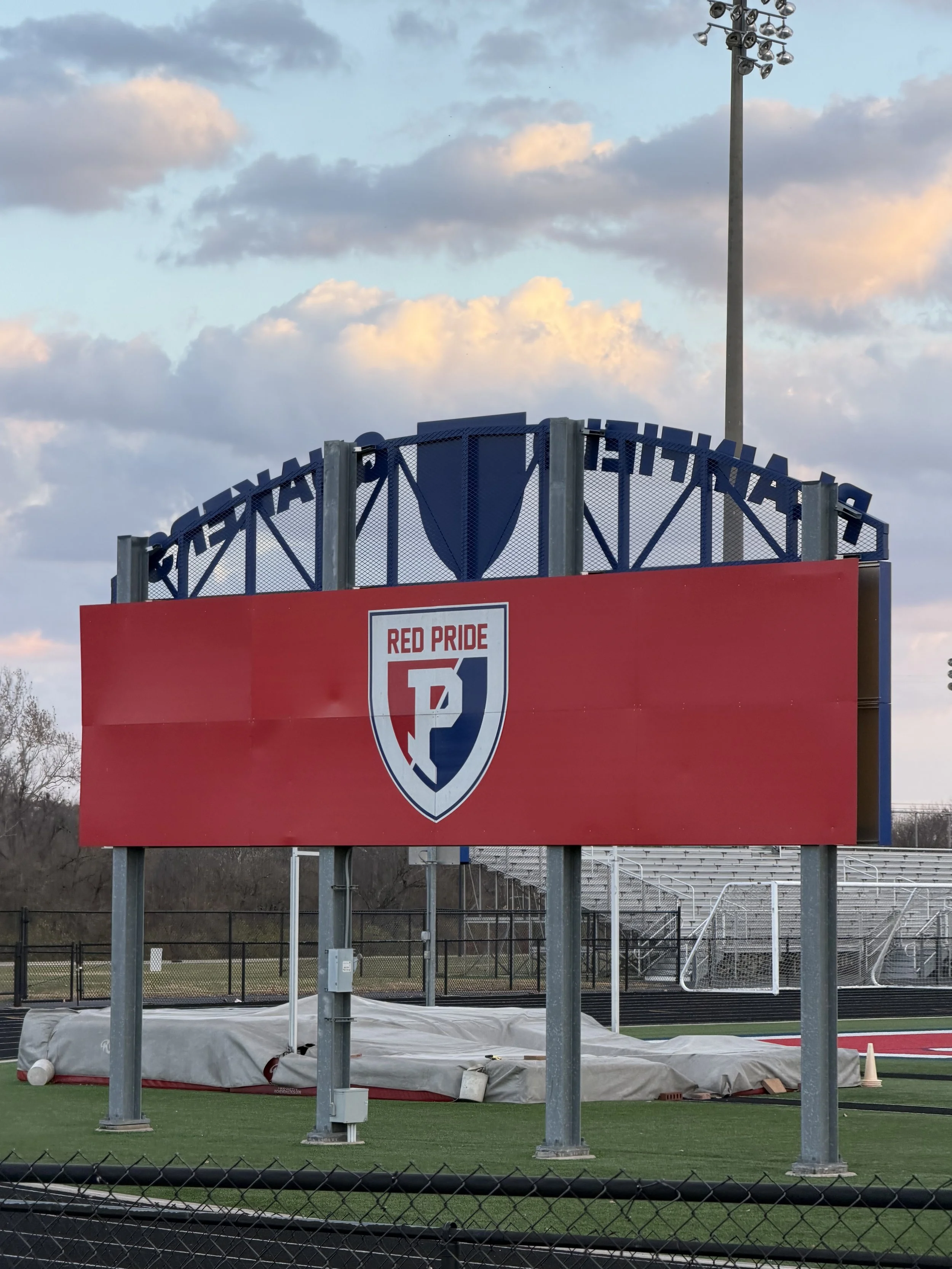A sports scoreboard at a football field displays a logo for Red Pride, with a goalpost and stadium seating in the background, under a partly cloudy sky.
