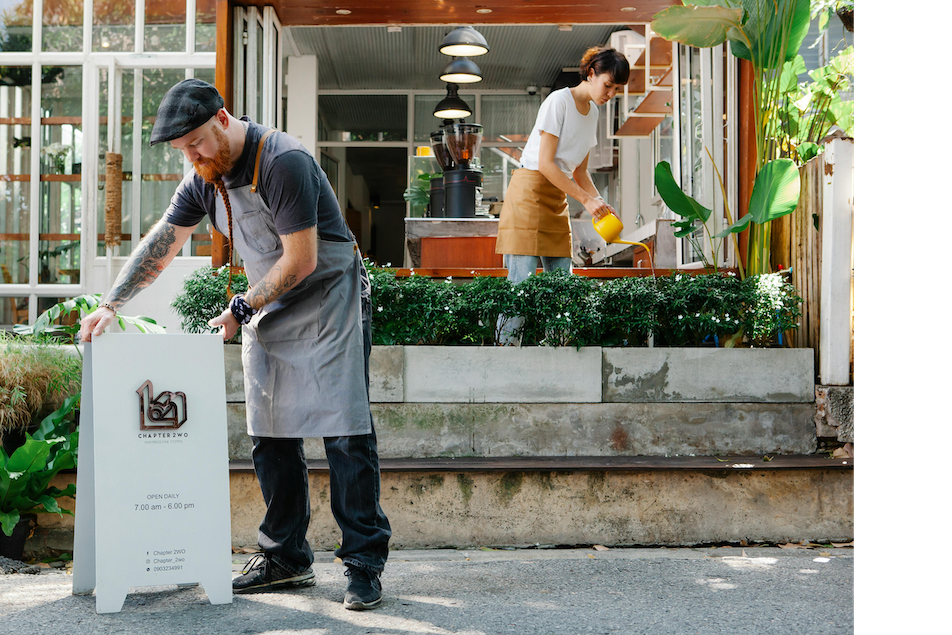 A man and a woman working outside a cafe or restaurant. The man is cleaning a signboard, and the woman is watering plants on a raised garden bed.