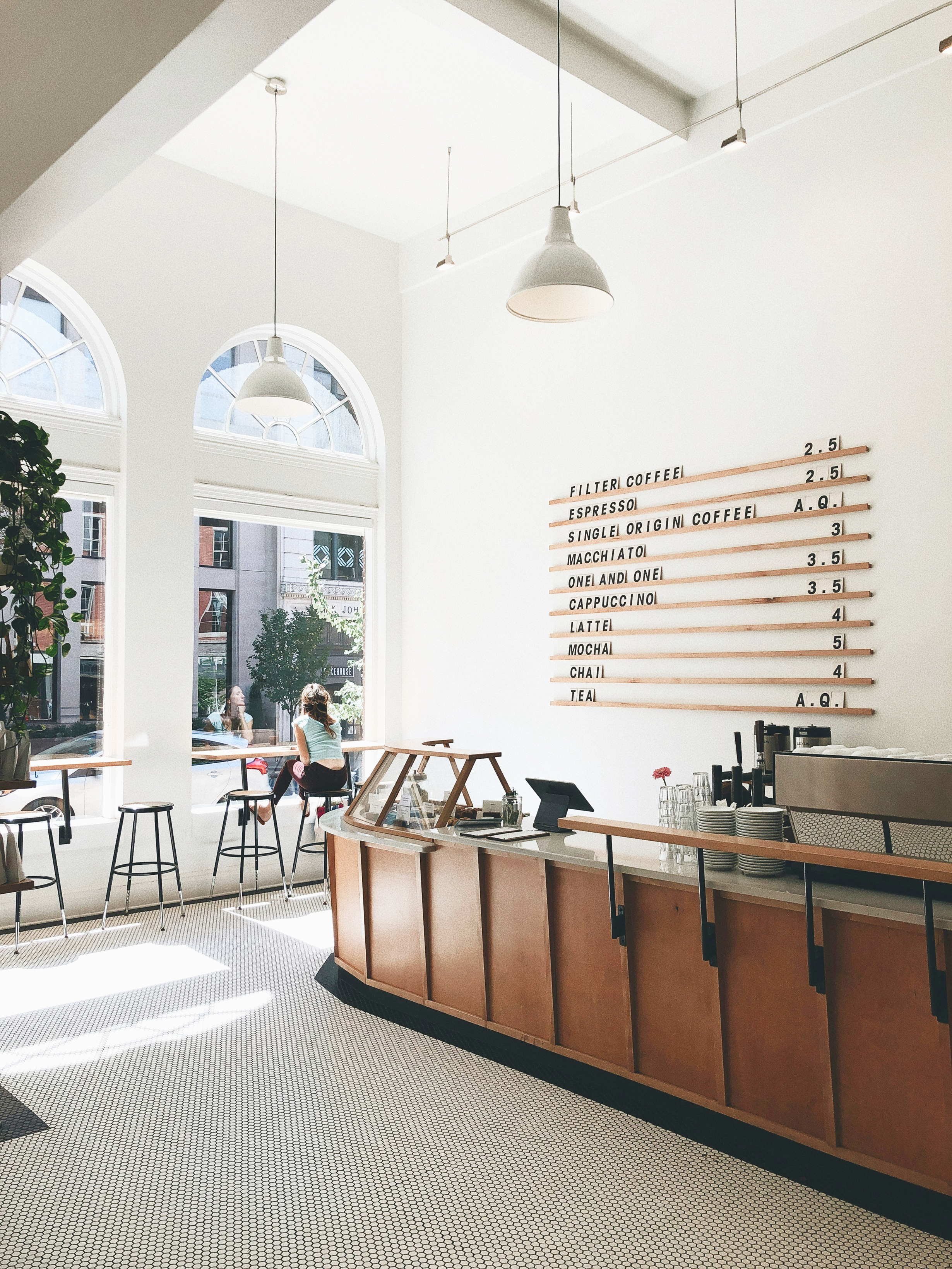 Interior of a bright coffee shop with large arched windows, a wooden counter, and a menu board on the wall displaying coffee options and prices. Two women are sitting by the window.