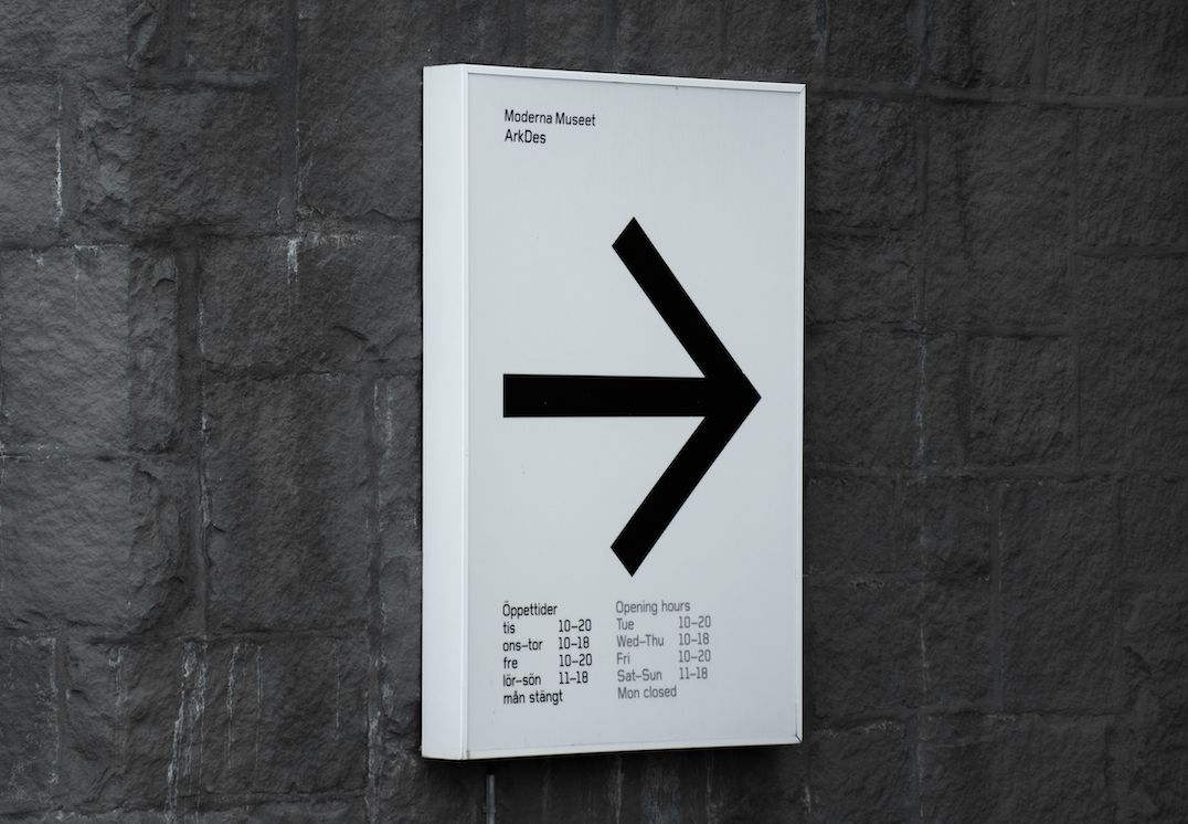 A white sign with black text and a large black arrow pointing to the right, mounted on a gray brick wall.