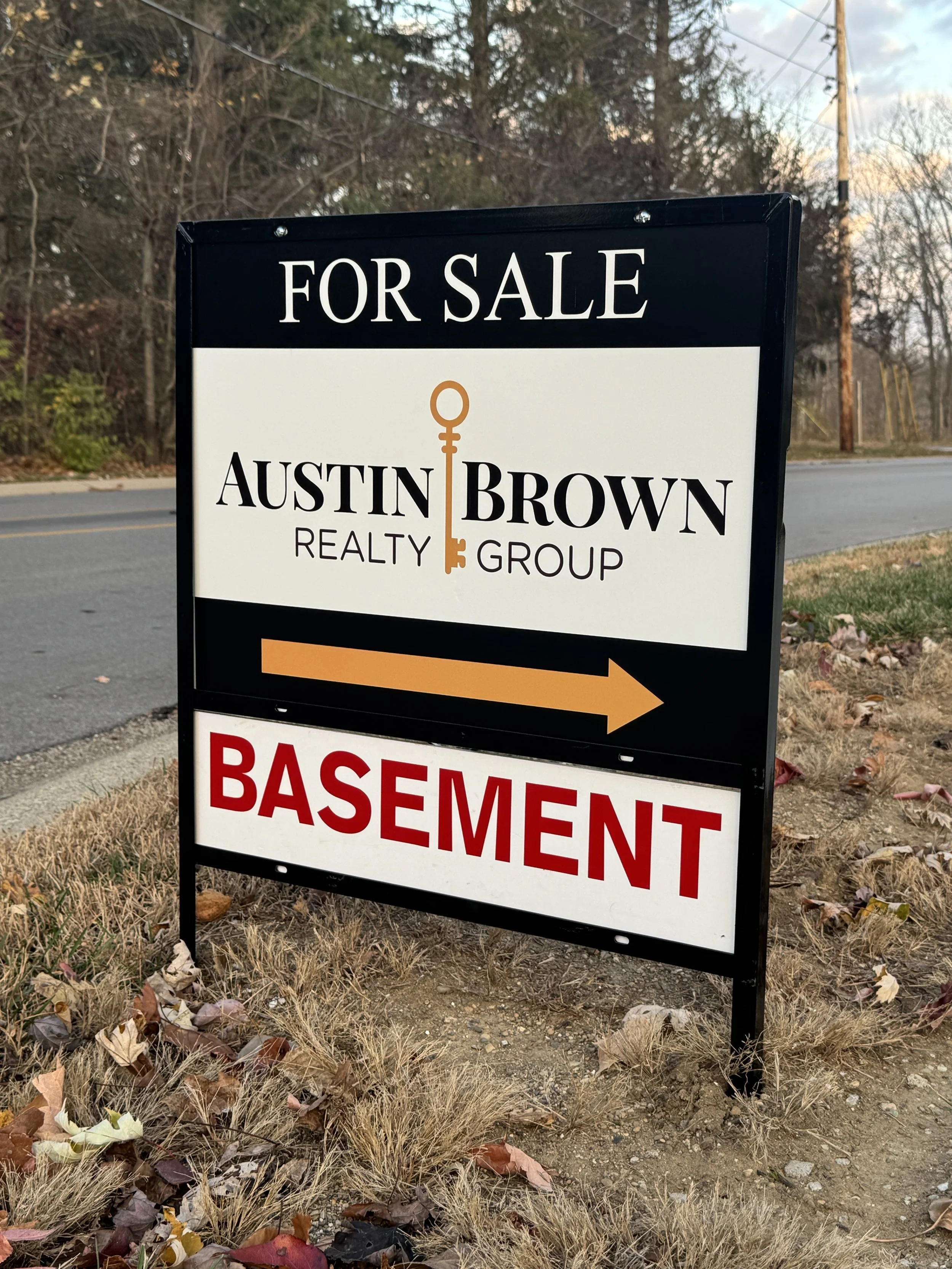 A driveway sign with black and white background, advertising for sale by Austin Brown Realty Group including directional arrow pointing right to a basement.