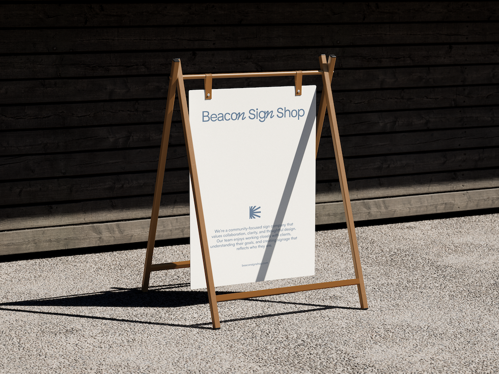 A white A-frame sidewalk sign with the words 'Beacon Sign Shop' at the top, placed on a light-colored gravel surface in front of a dark wooden wall, with text and logo on the sign.