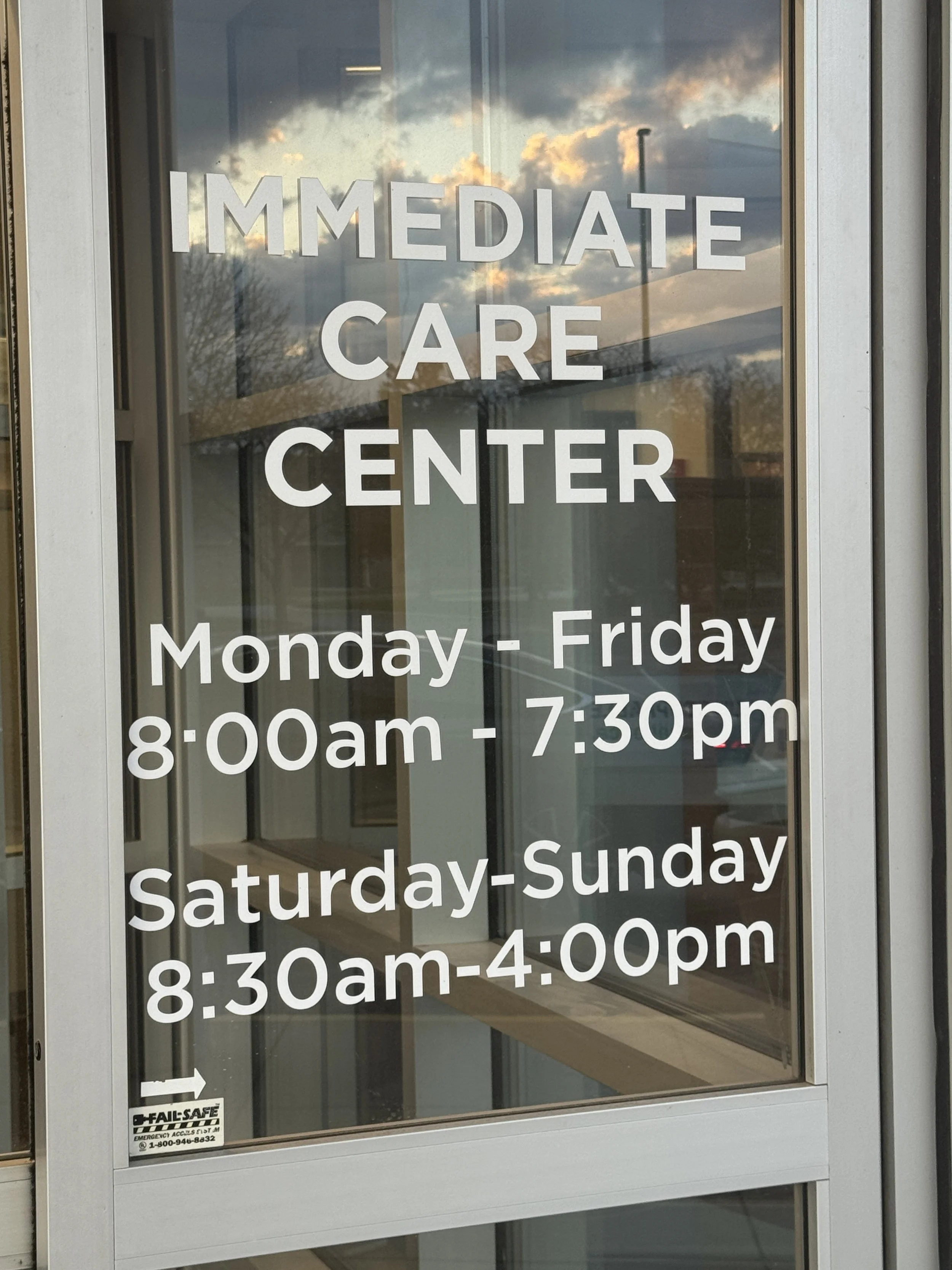 Glass door with sign stating hours for an immediate care center, open weekdays from 8:00 am to 7:30 pm and weekends from 8:30 am to 4:00 pm, with a reflection of the sky and clouds nearby.