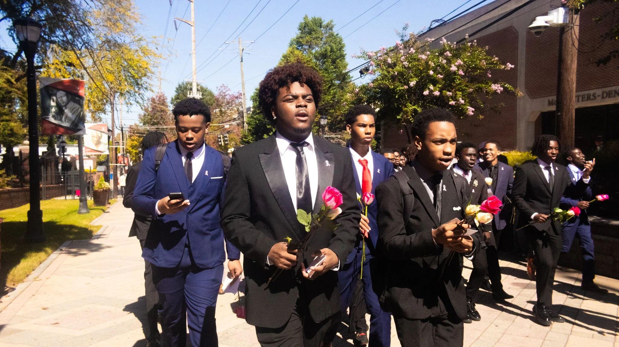 A group of young men dressed in suits participating in a memorial march, holding roses and walking on a city sidewalk.