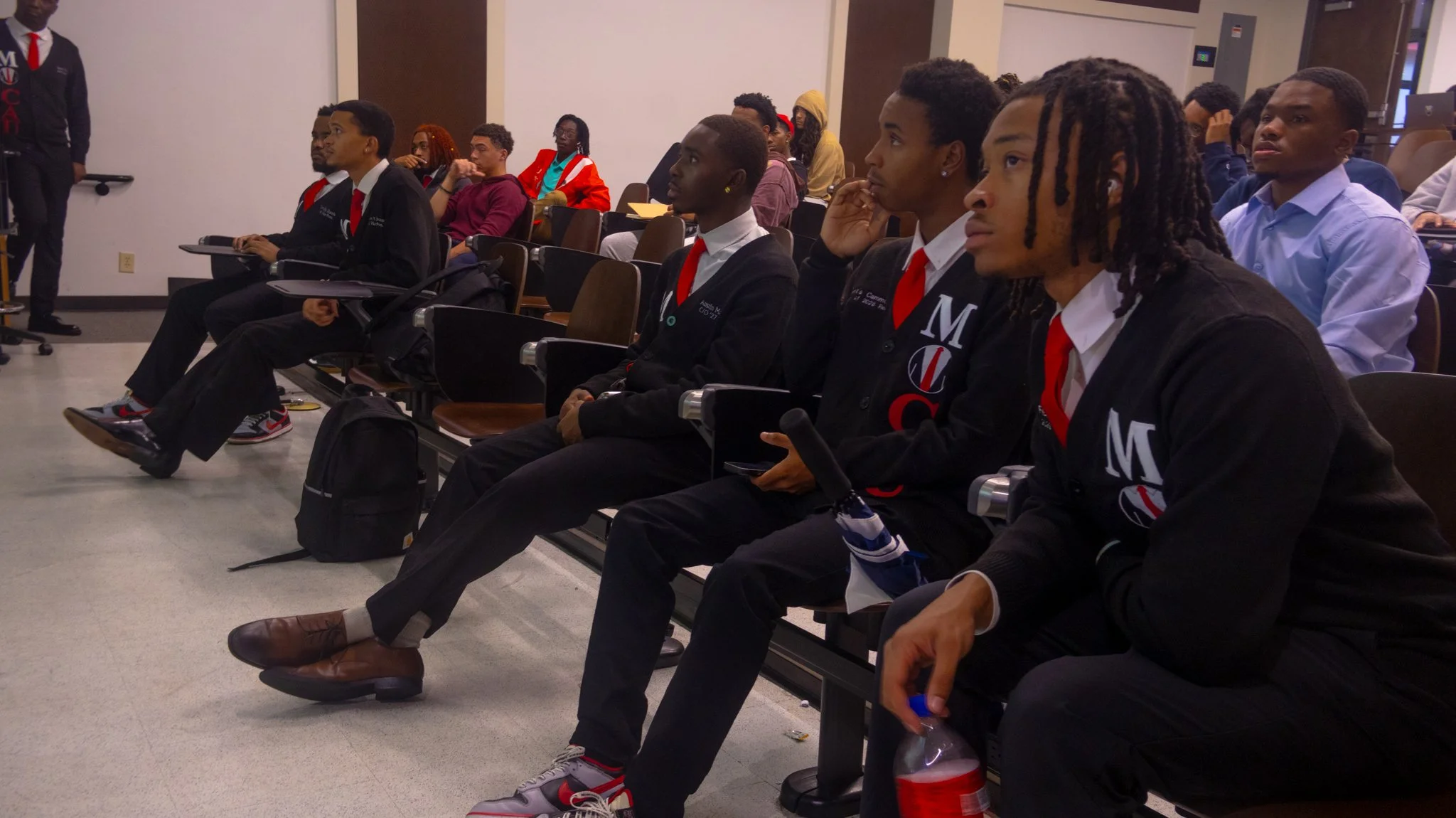 Young men and women in formal attire seated in an auditorium, listening attentively, with some holding notebooks or bottled drinks.