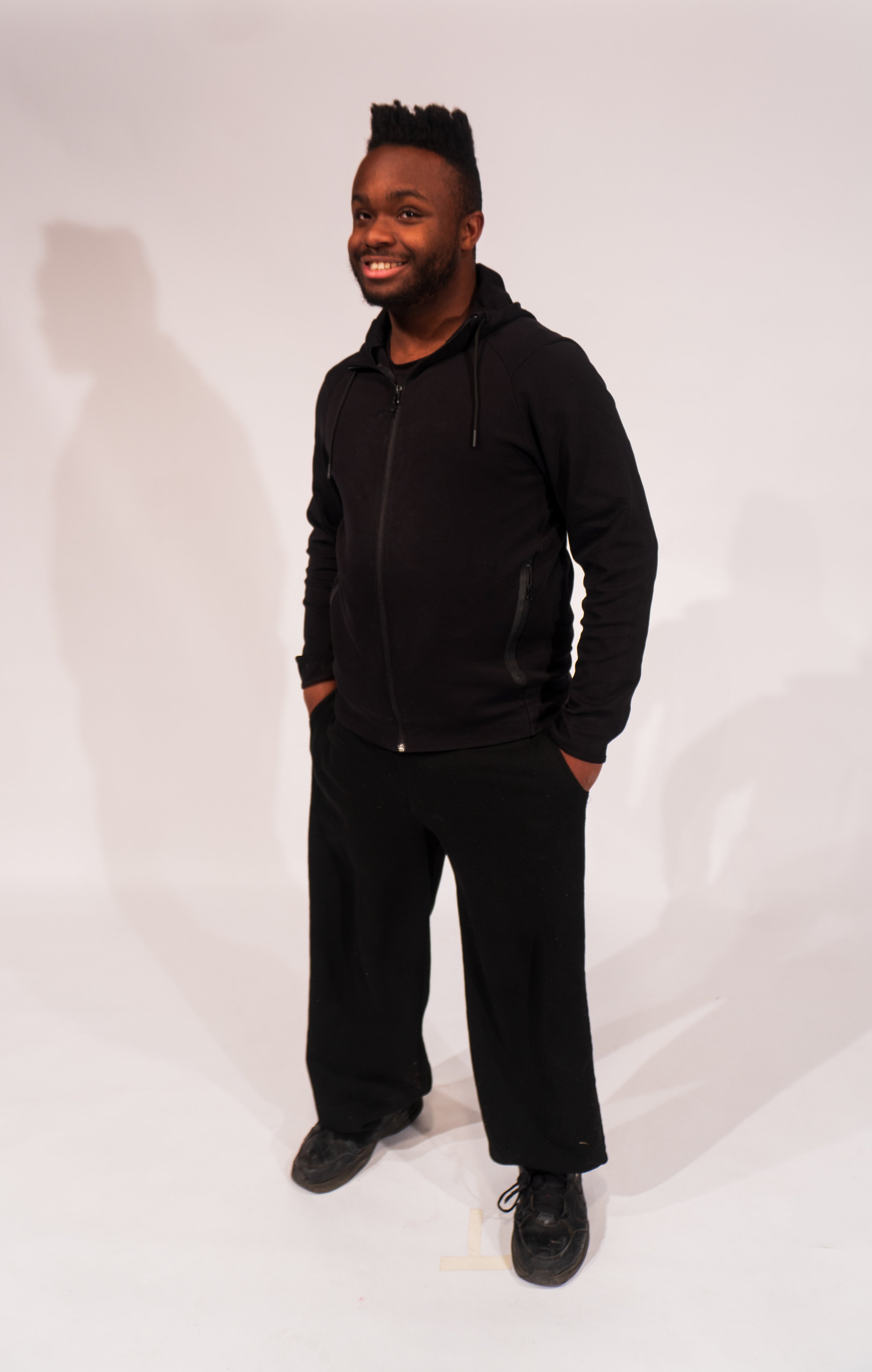 A smiling man with dark skin, sporting a high-top hairstyle, wearing a black zip-up hoodie, black pants, and black shoes, standing against a plain white background with his hands in his pockets.