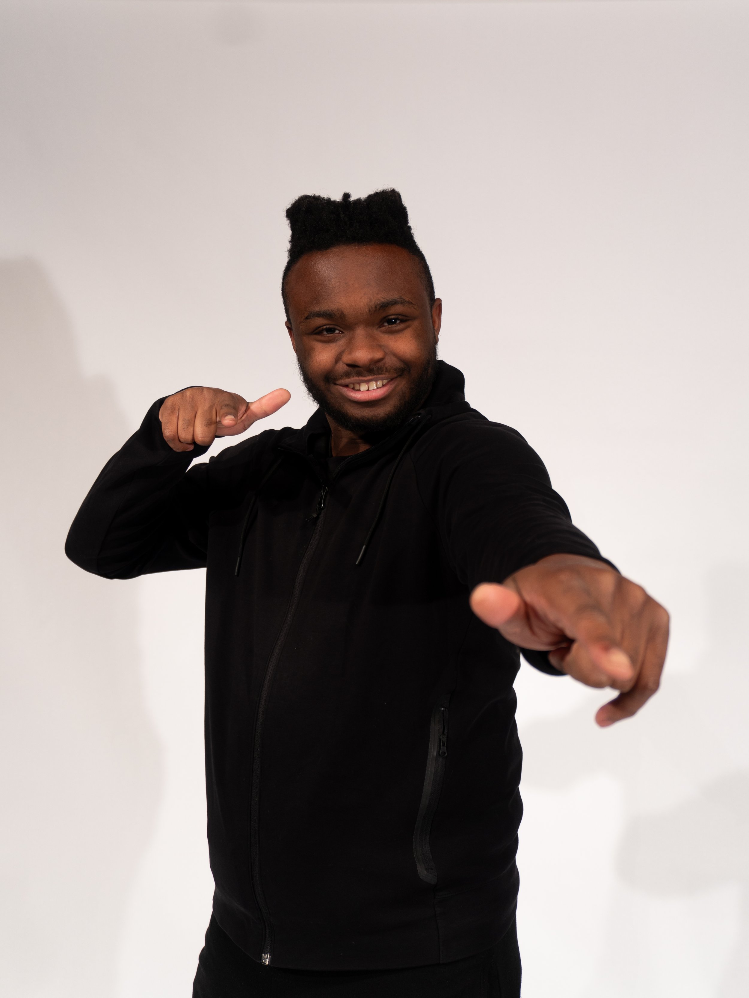 A smiling man with dark skin and styled hair pointing and gesturing towards the camera against a plain white background.