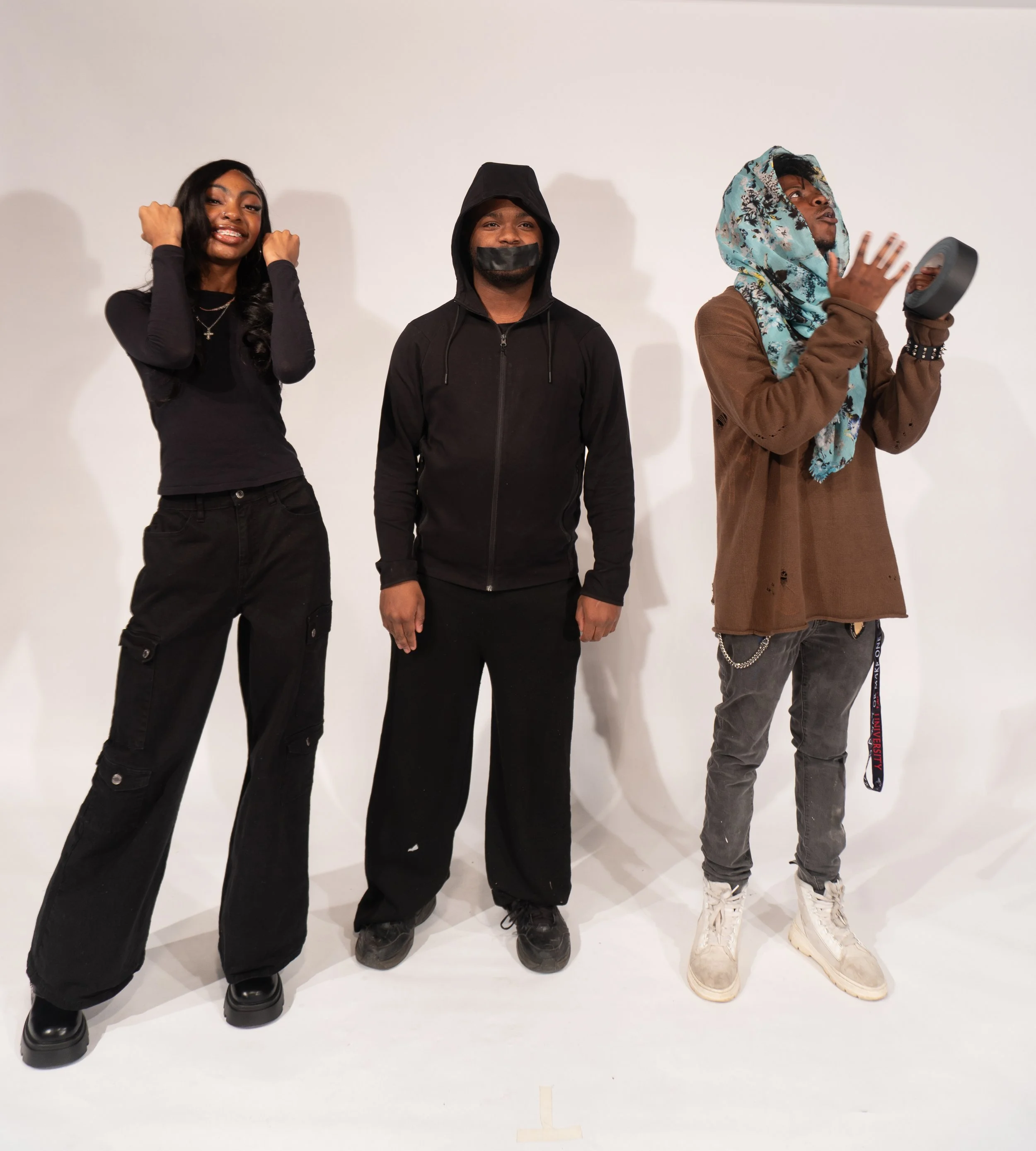 Three young people standing against a plain white wall, dressed in casual streetwear, with one person wearing a face mask, and another holding a suncast tape roll.