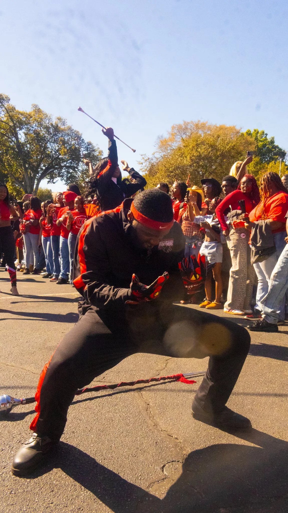 A man dressed in black sportswear with red accents dancing or performing in front of a group of people, some wearing red apparel, during an outdoor event or parade. The crowd is taking pictures and watching, with trees in the background and a clear blue sky.