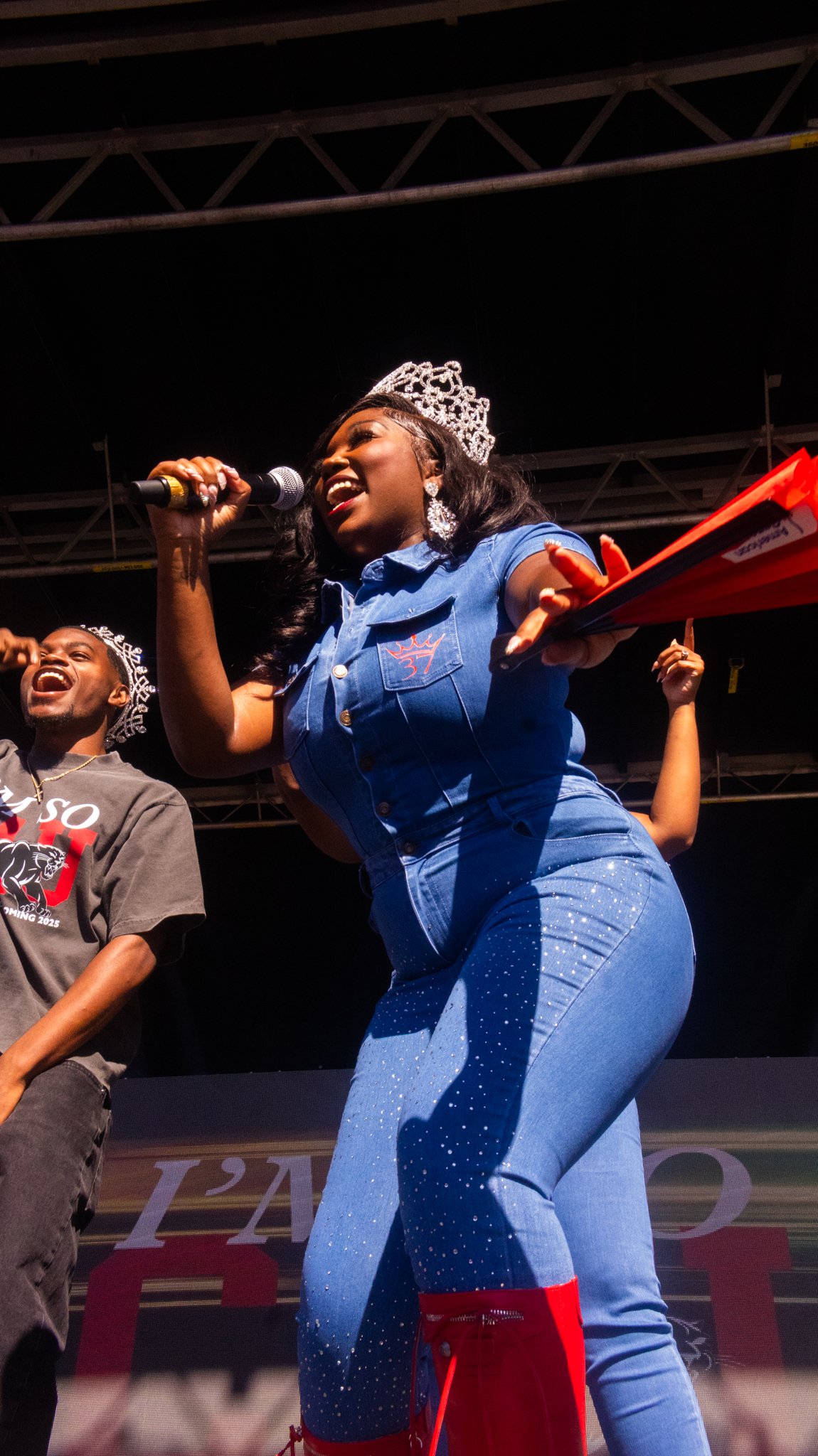 A woman wearing a crown, earrings, and a blue outfit with sparkling details is speaking into a microphone on stage. A man beside her is smiling and wearing a crown and a gray T-shirt. The stage has a black backdrop and metal framework overhead.