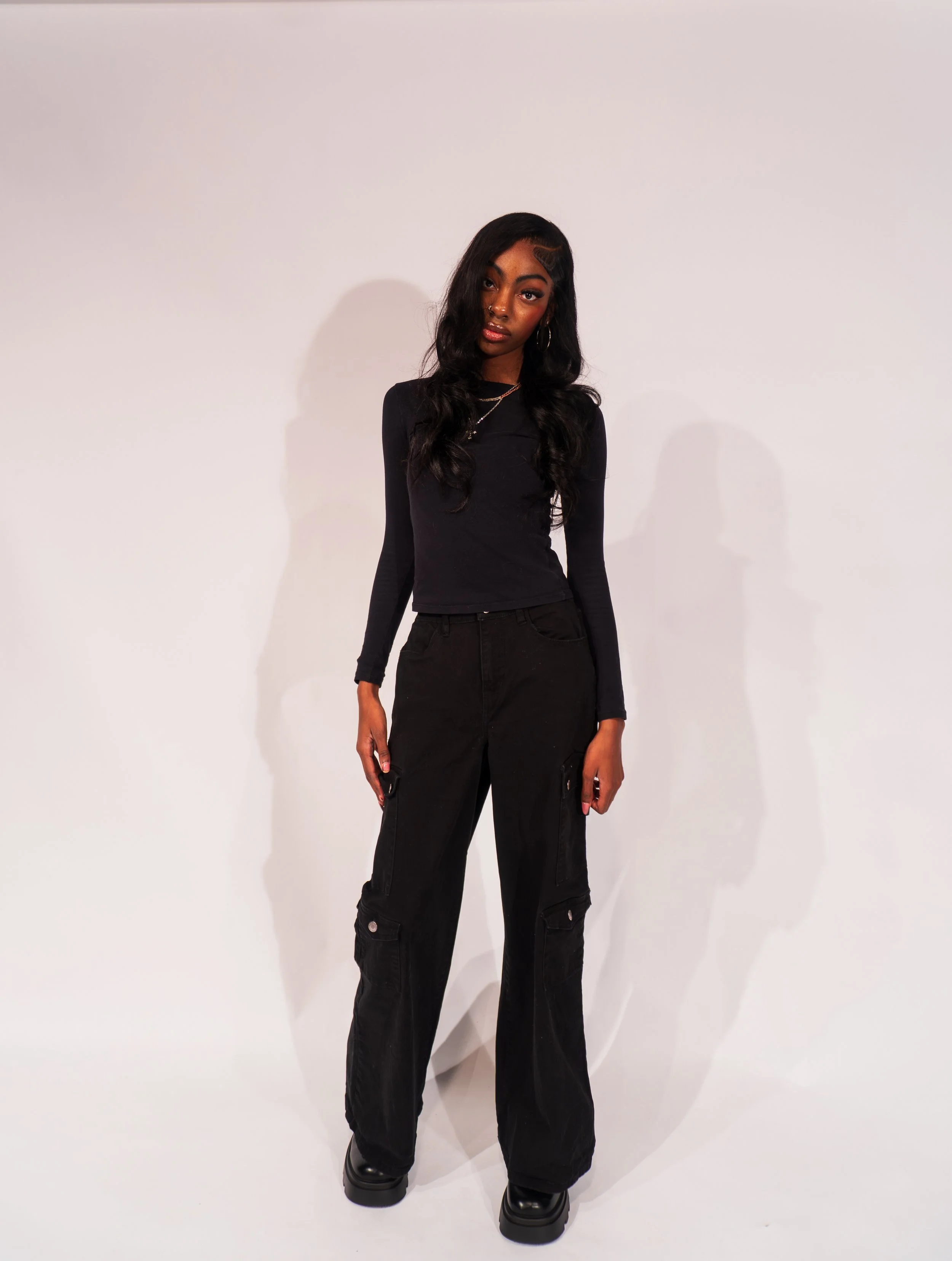 Young woman with dark hair in loose waves, wearing a fitted black long-sleeve top, black cargo pants, and black boots, standing against a plain white background.