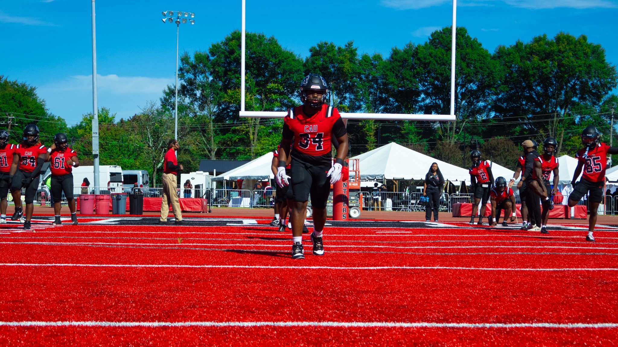 Football players in red and black uniforms warming up on a red track at an outdoor sports field with white tents and tall trees in the background.