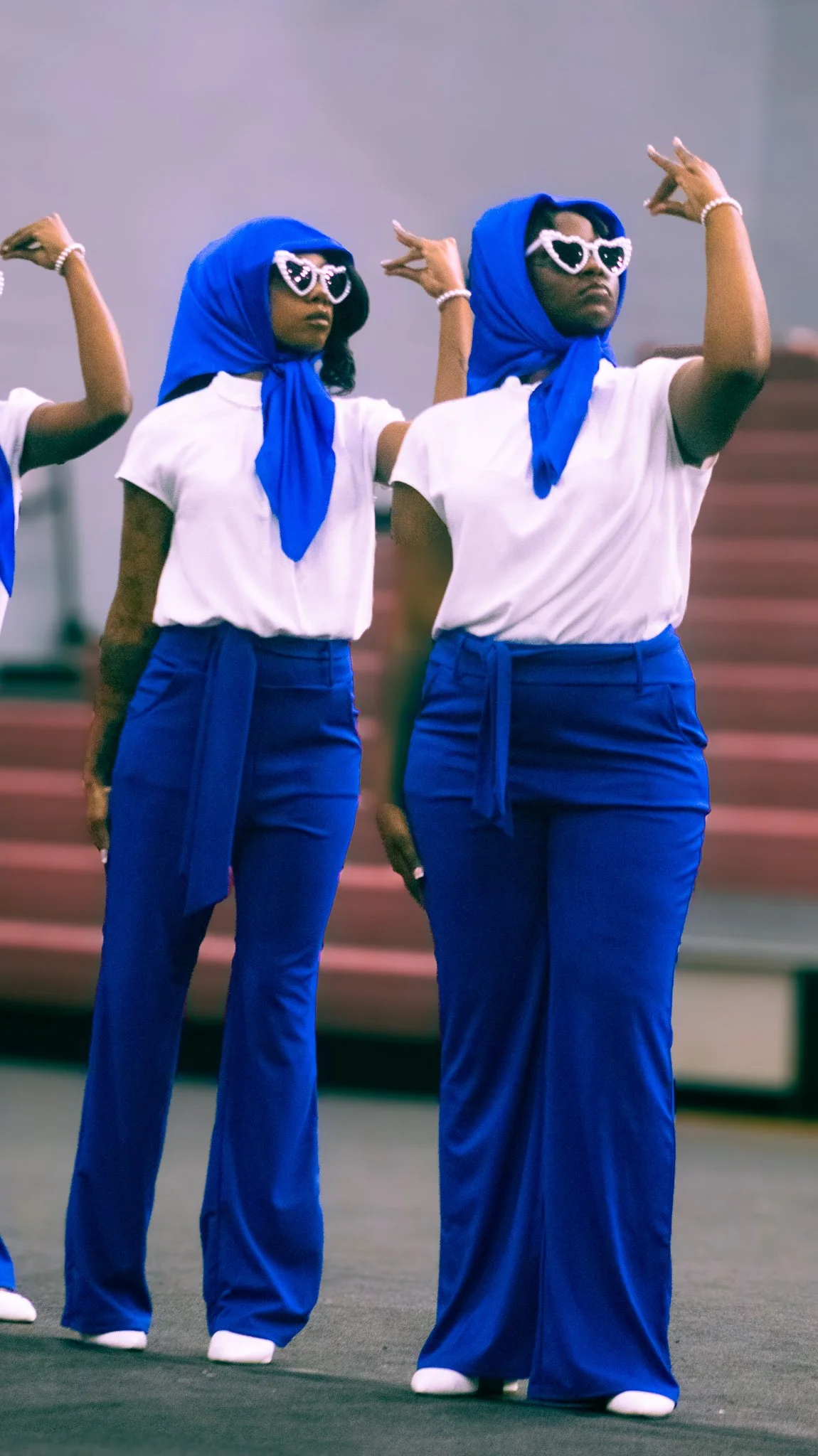 Women dressed in white T-shirts, blue headscarves, and blue wide-leg pants wearing heart-shaped sunglasses and pearl bracelets performing a dance move indoors.
