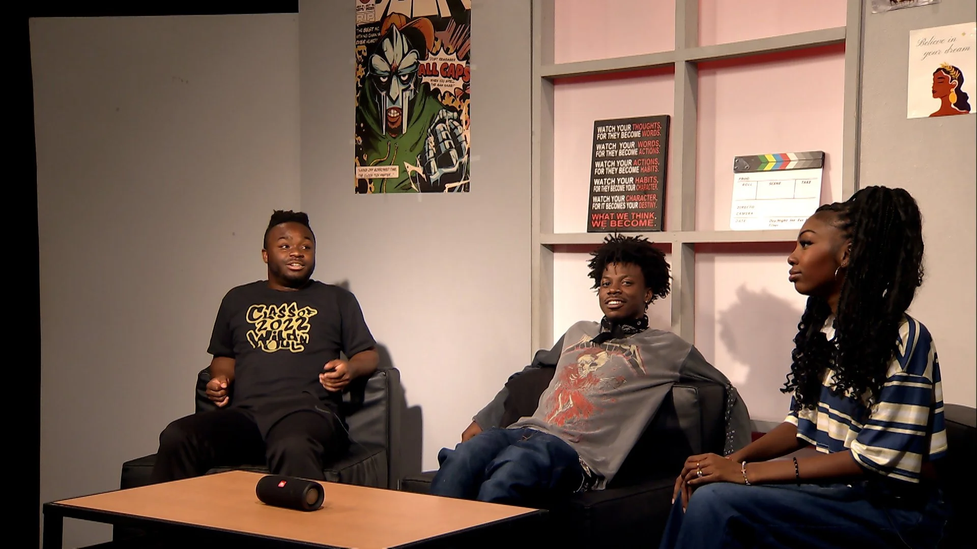 Three young adults sitting on couches in a room with posters on the wall, having a conversation. One is a man in a black t-shirt, another is a man with curly hair wearing a gray shirt, and the third is a woman with long braids wearing a striped top.