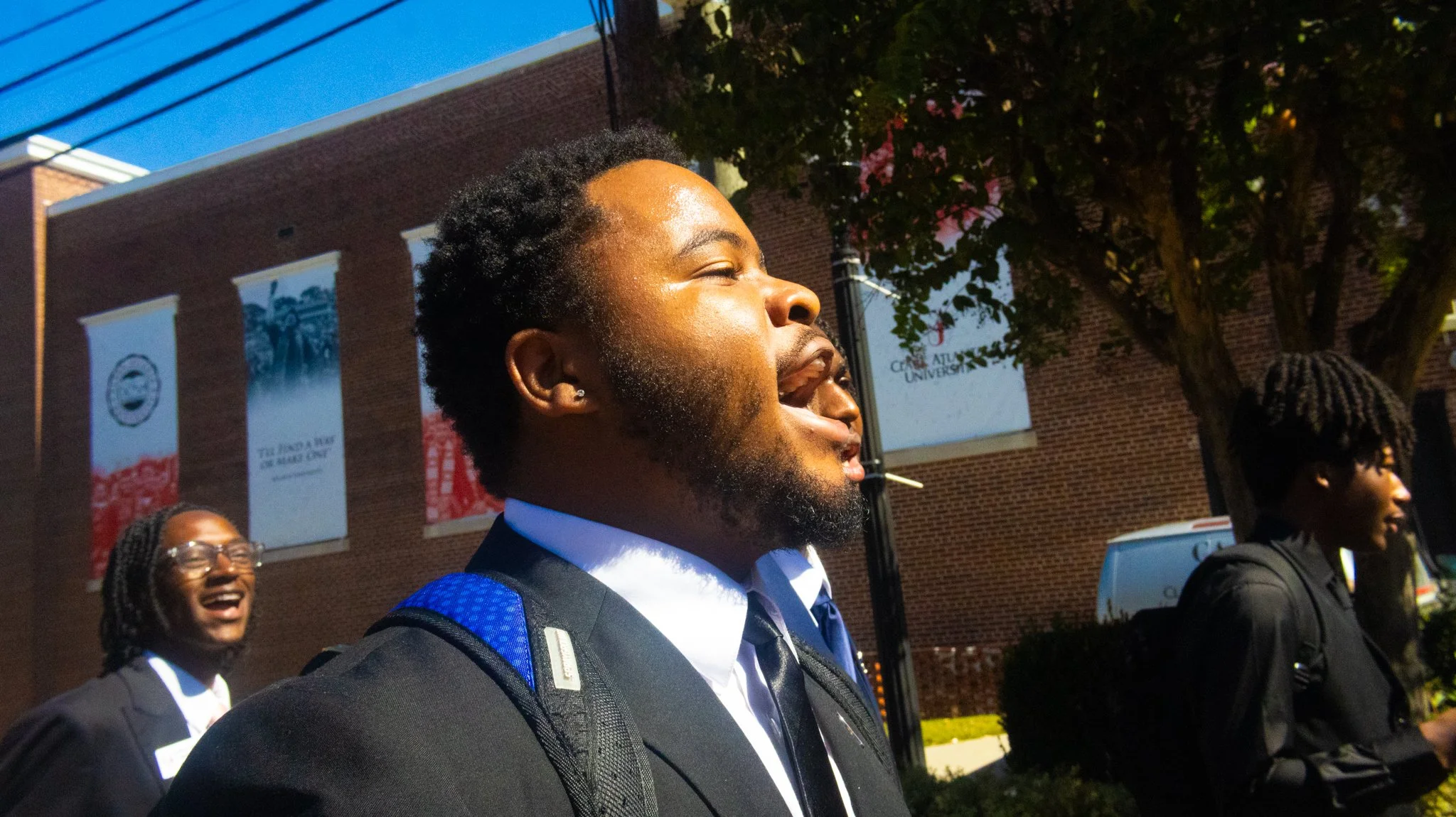 Group of students in suits standing outside a university building with banners, one student in the foreground is smiling with eyes closed, wearing a black suit and backpack, another is laughing and wearing glasses, and a third student with dreadlocks also in formal attire.