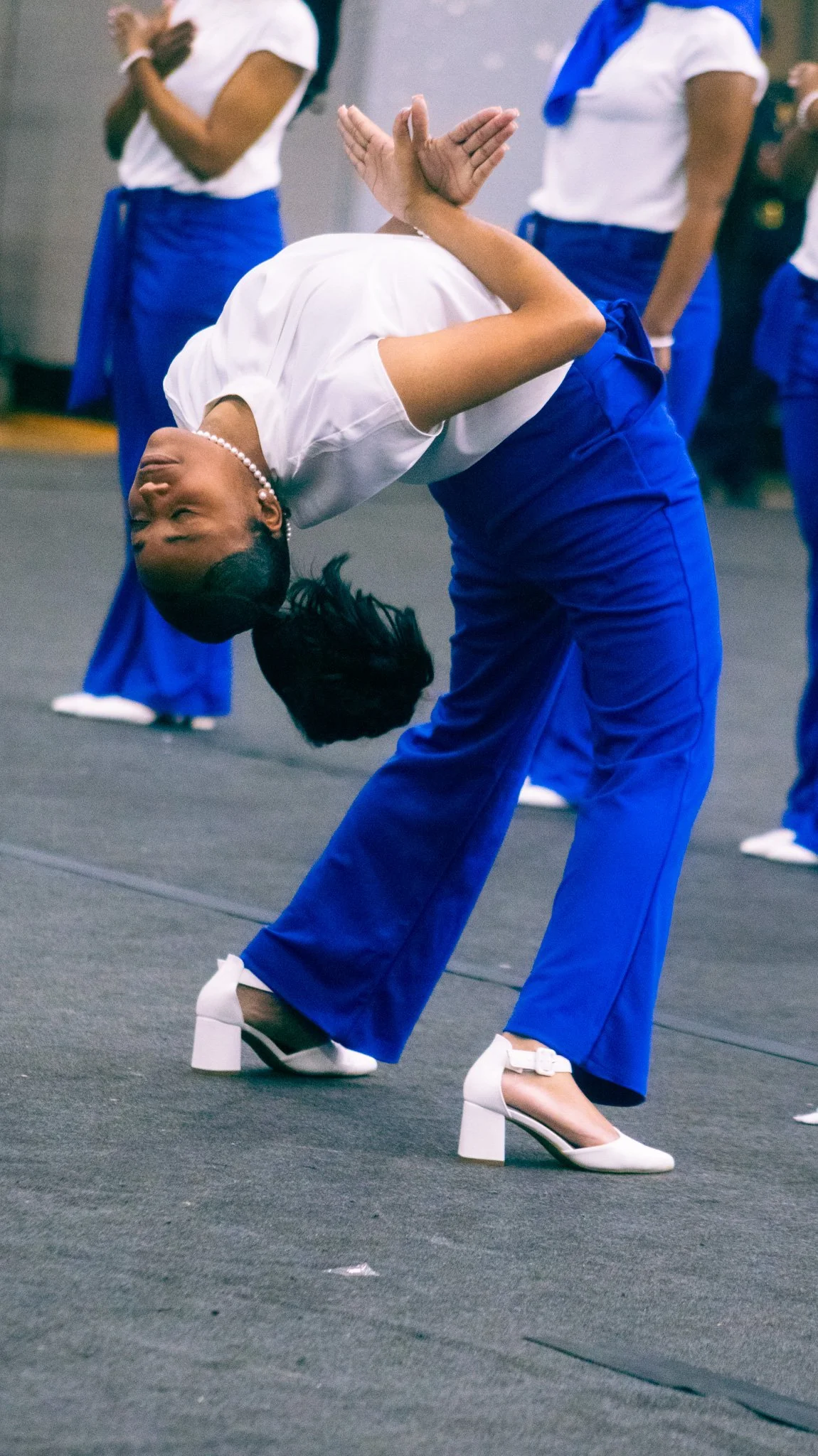 Woman wearing blue pants and white high heels dancing with her arm raised and head tilted back, surrounded by other women in similar attire, in a room with a dark floor.