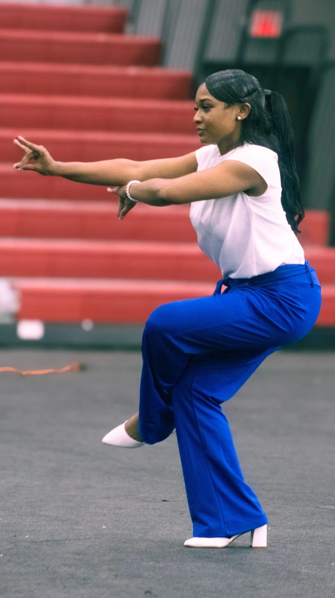 A woman in blue pants, a white top, and white heels is performing a dance move, balancing on one leg in an indoor gym with red bleacher seats in the background.