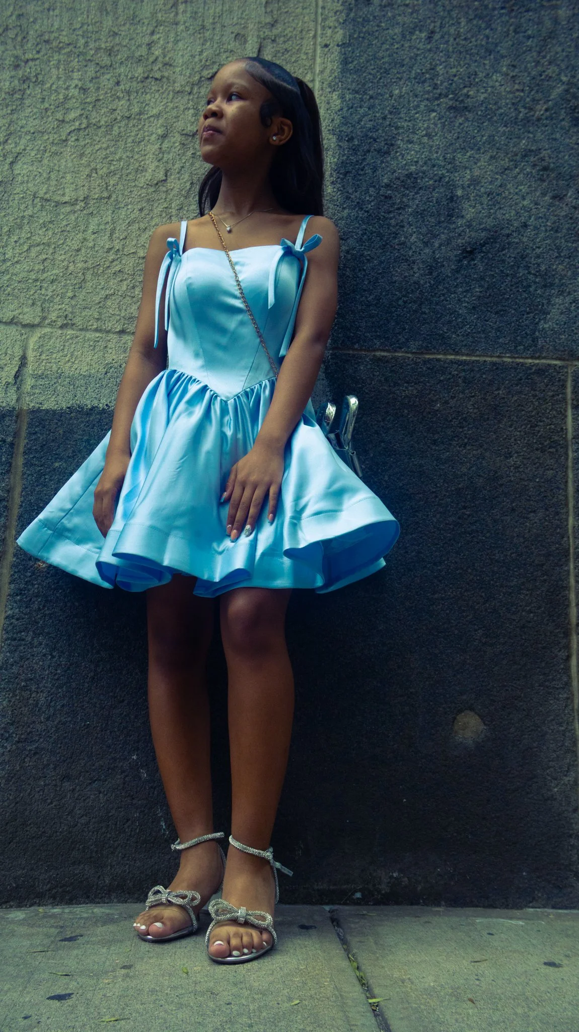 Young girl in a shiny blue dress with bow details, wearing high-heeled sandals with bows, standing against a textured wall.