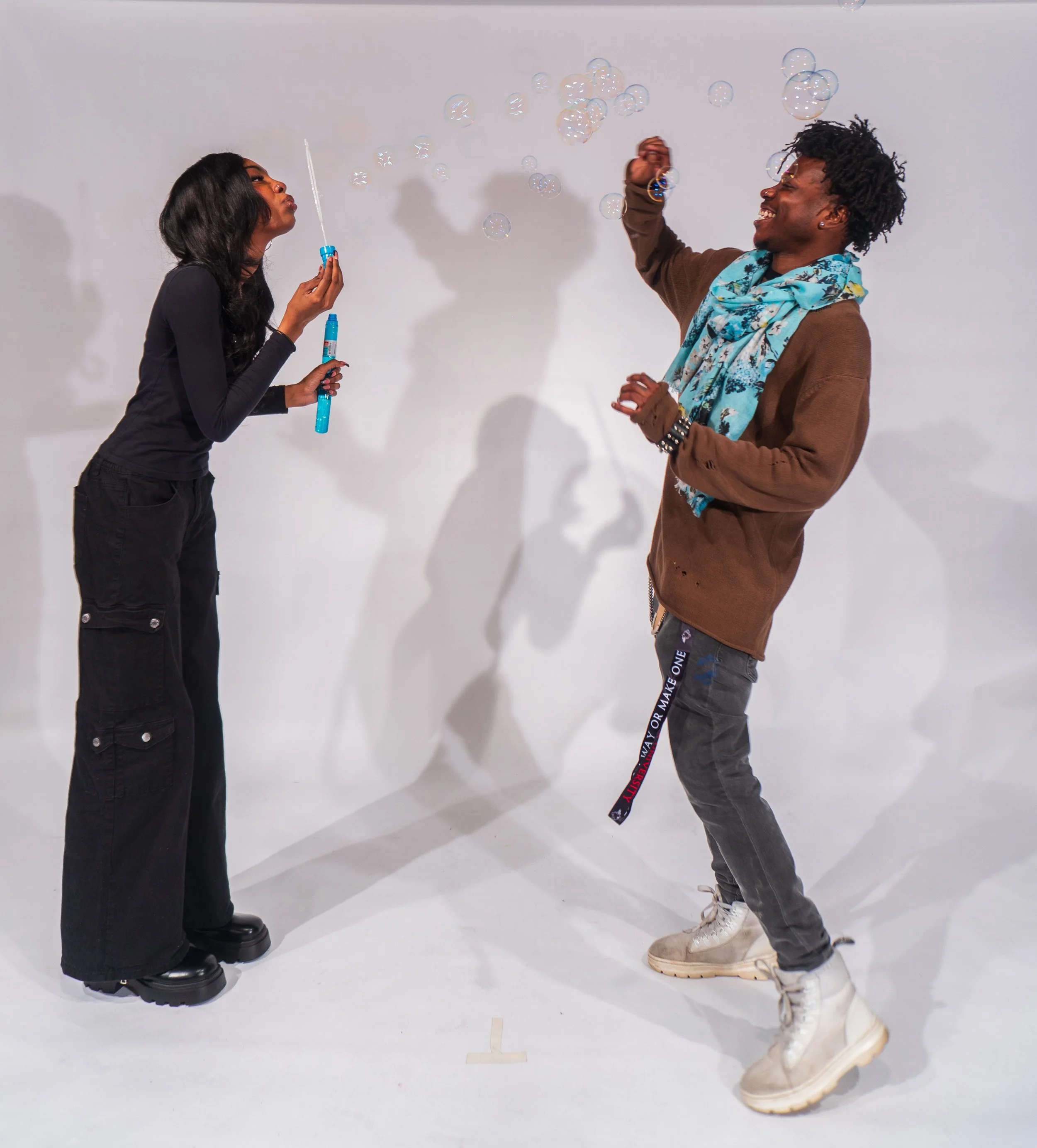 A woman and a man are playing with soap bubbles against a plain white background. The woman is holding a bubble wand and blowing bubbles, while the man is joyfully reaching toward the bubbles, wearing a brown jacket, a colorful scarf, and white boots.