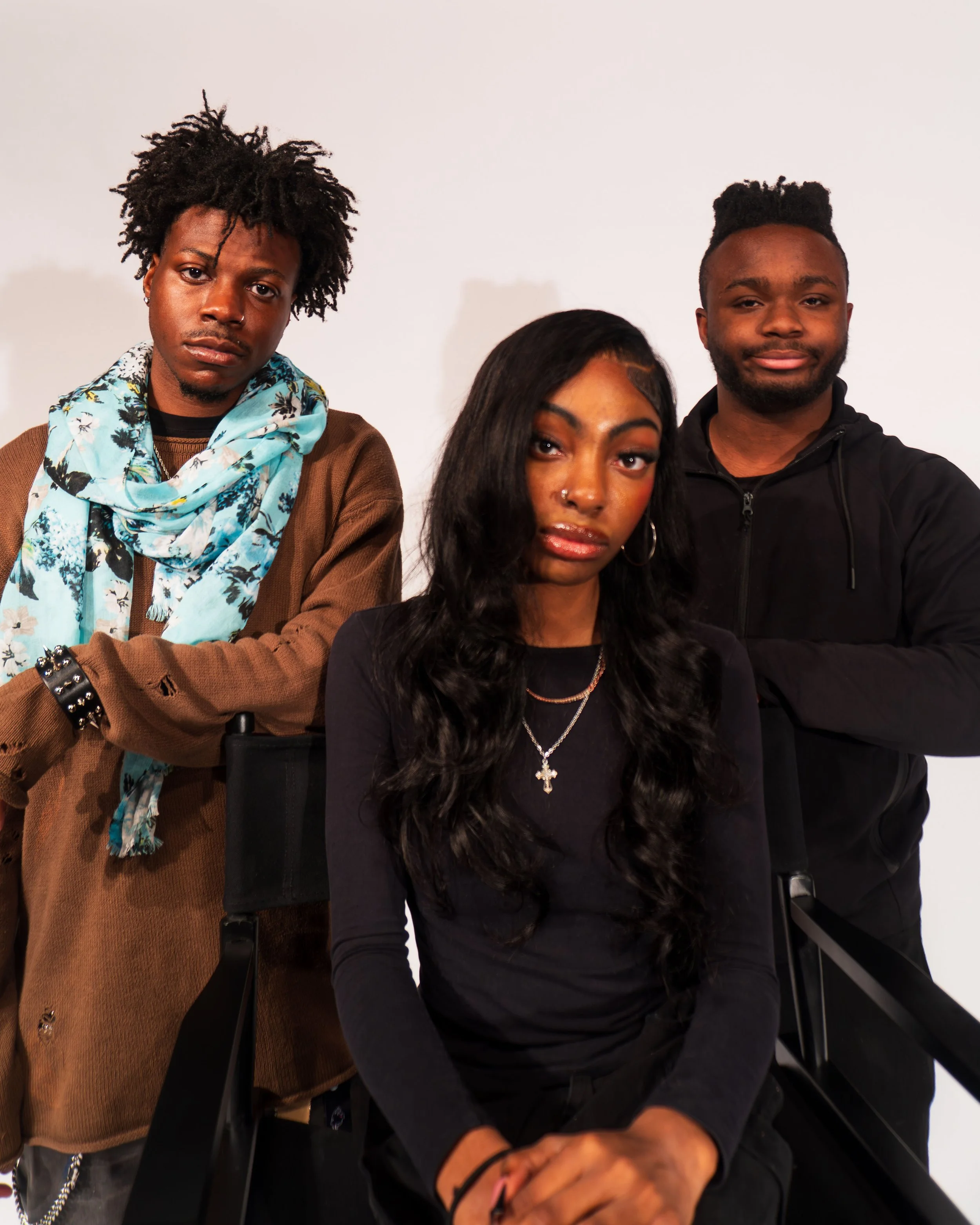 Three young adults pose for a photo in front of a plain white background. The woman in the center has long dark wavy hair, wears hoop earrings, a black shirt, and layered necklaces. The man on the left has afro-textured hair, wears a brown shirt with ripped sleeves and a light blue patterned scarf, and has a serious expression. The man on the right has short curly hair, wears a black zip-up hoodie, and is smiling.